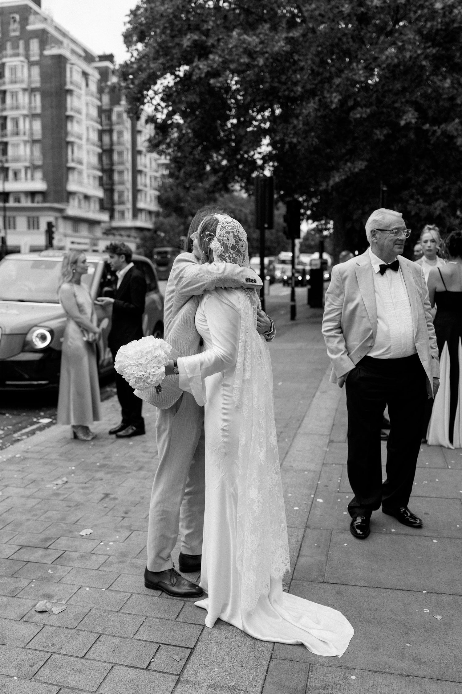 Black and white outdoor wedding photo showing embrace on city sidewalk with urban buildings.