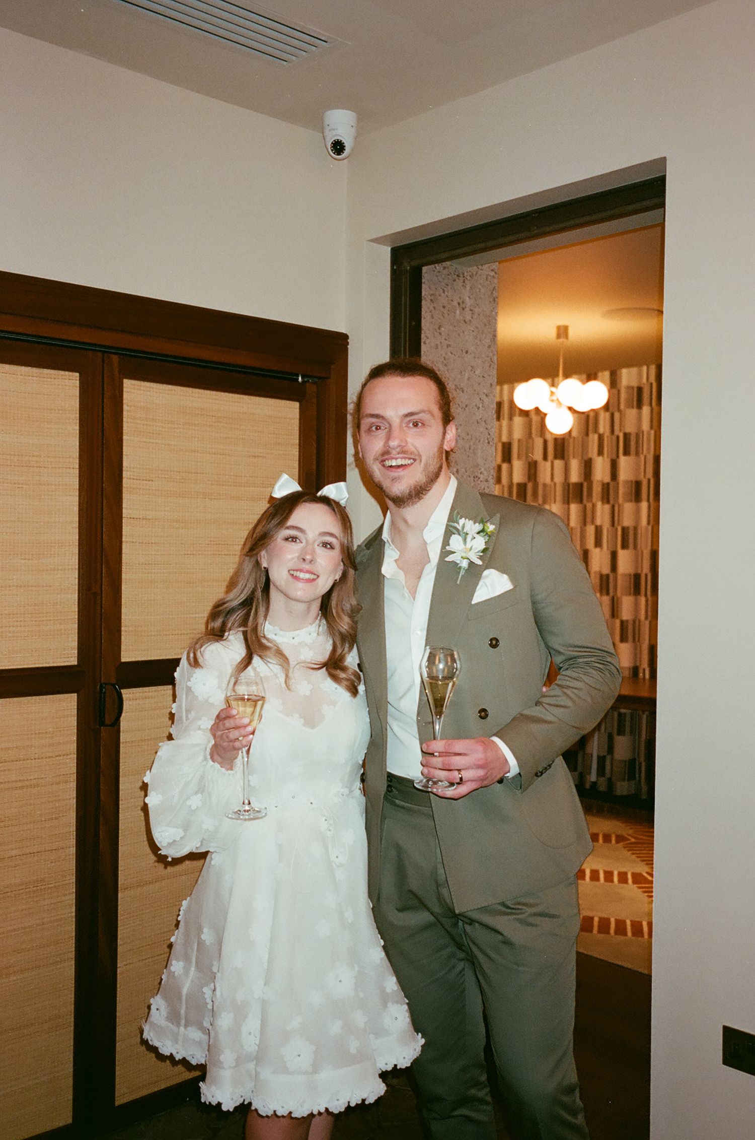 A couple in formal attire holds champagne glasses while posing together at an indoor celebration.