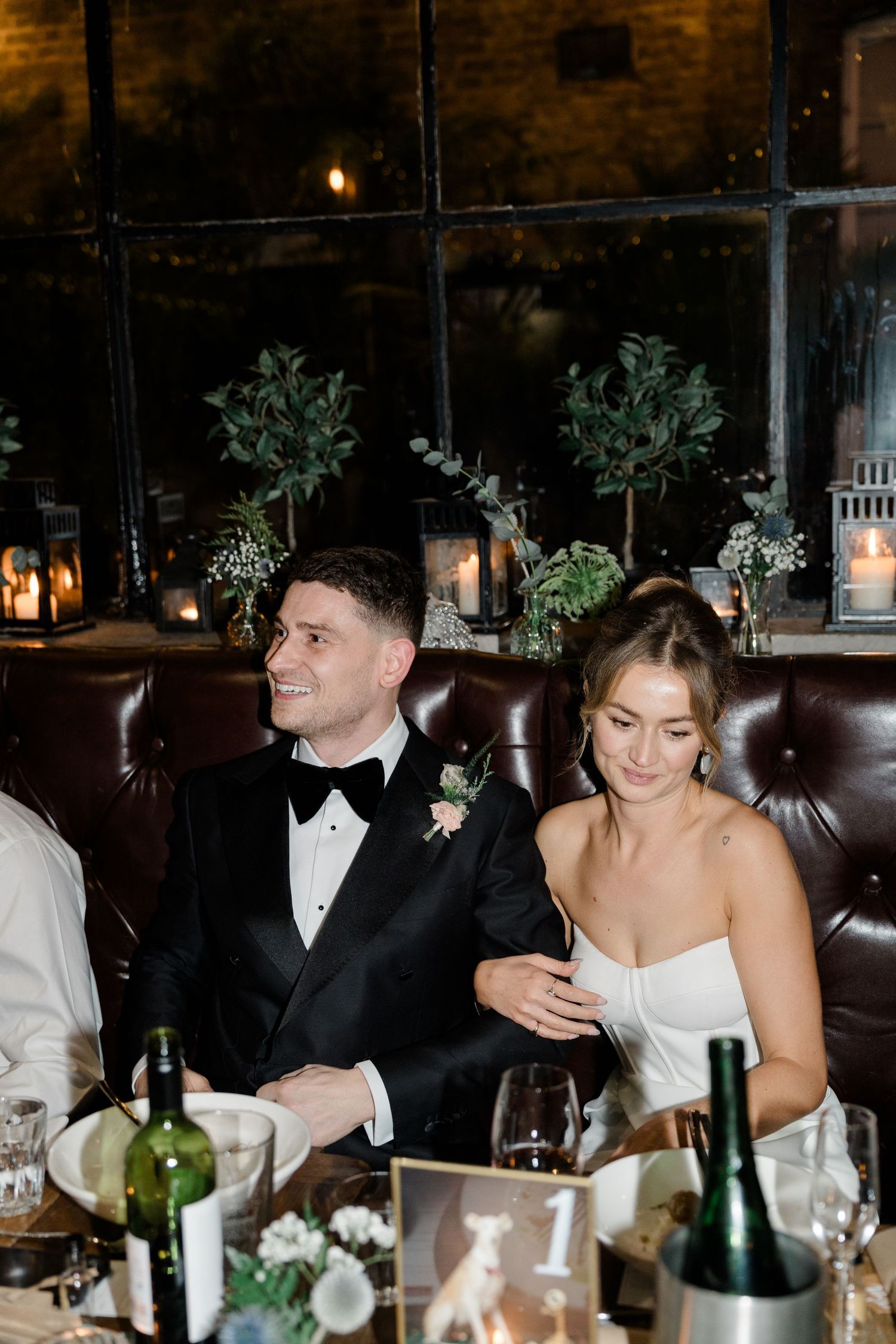 Wedding guests seated at a formal dinner table with wine bottles and decorative greenery in an elegant restaurant setting.