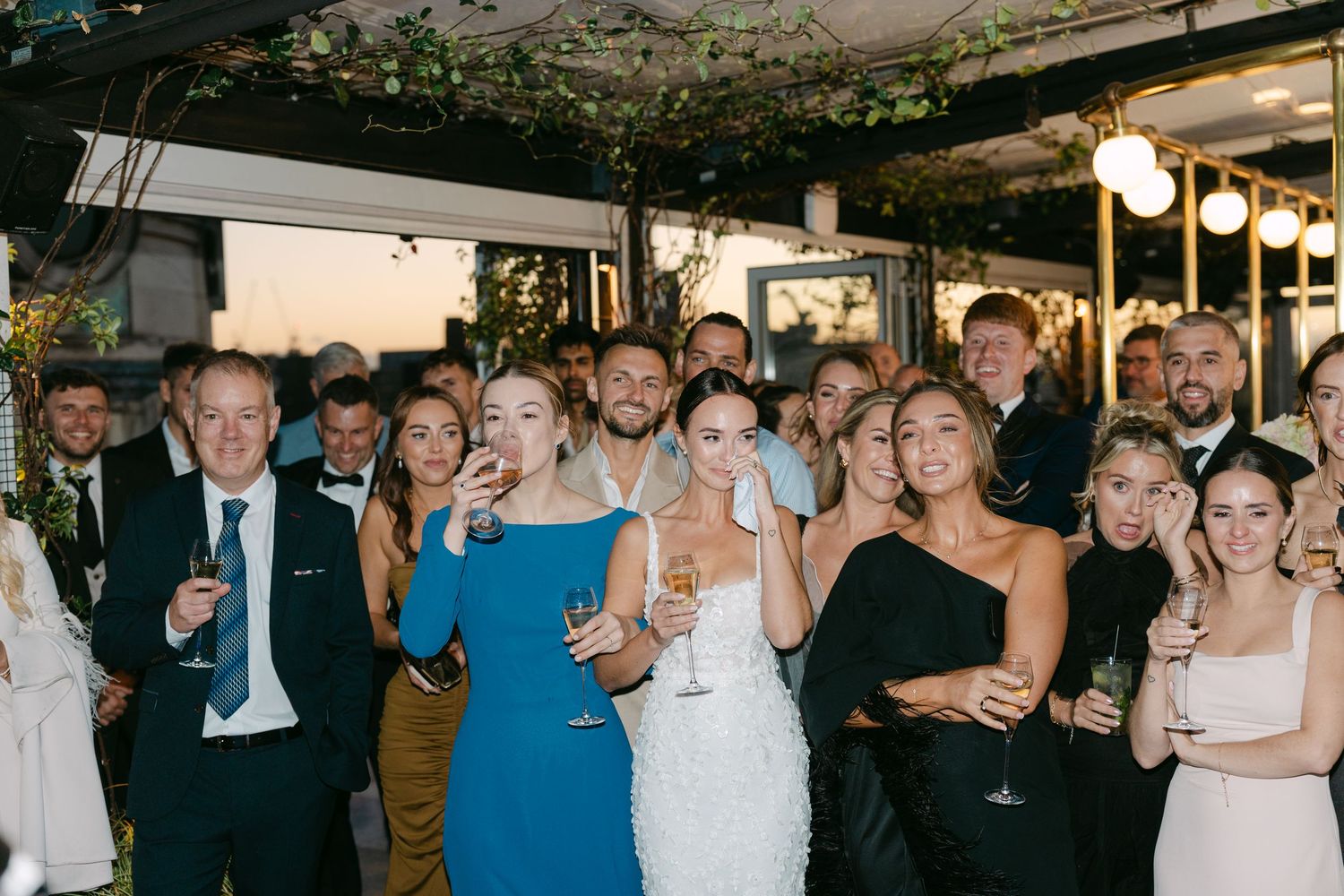 Wedding party and guests gather for a group moment under decorative ceiling greenery.