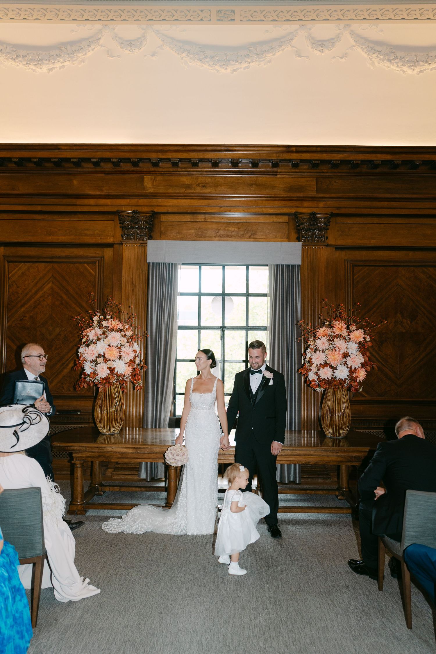 Wedding ceremony space with wooden paneled walls, floral arrangements, and seated guests.