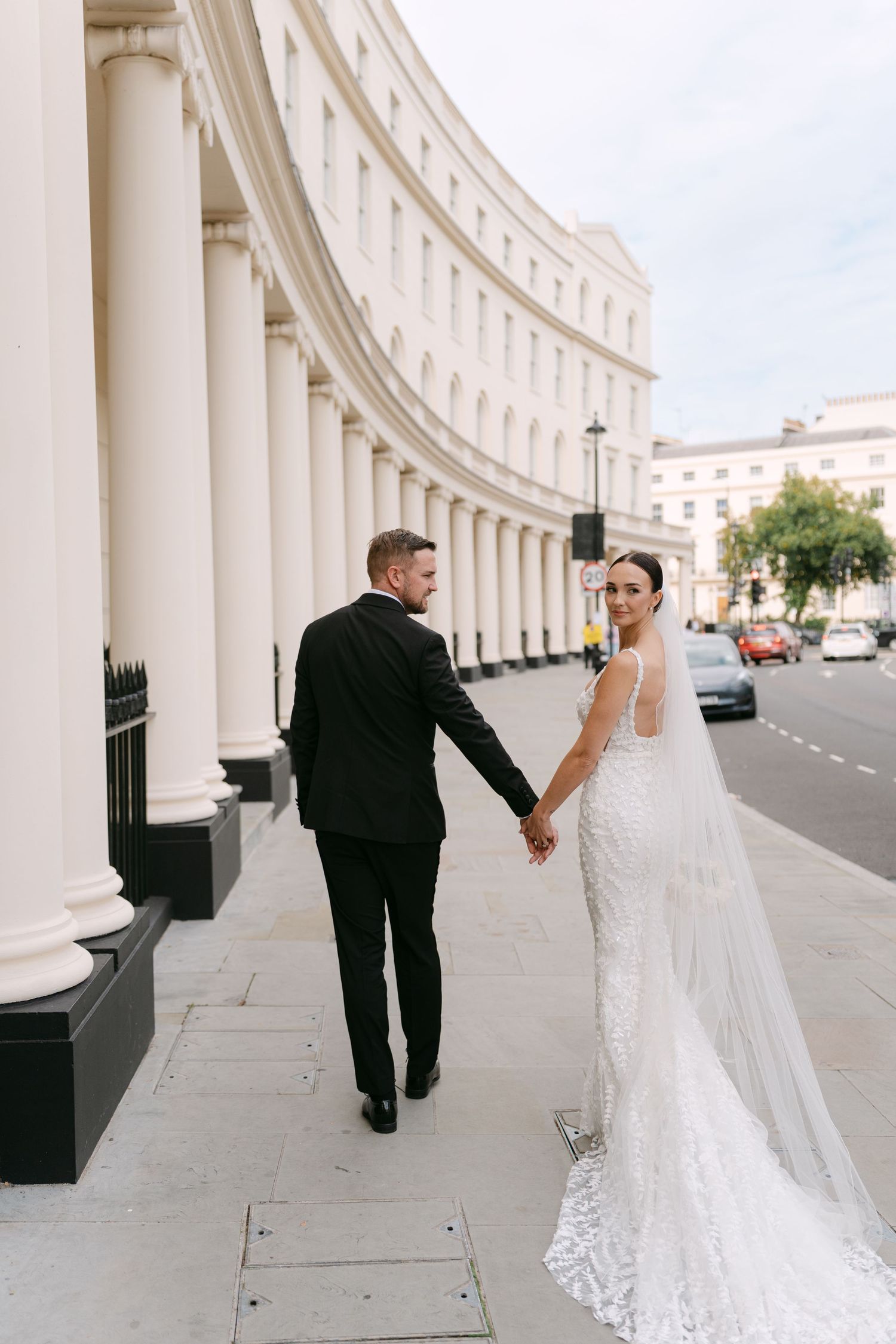 A bride in lace gown and groom walk hand in hand along a curved colonnade building.