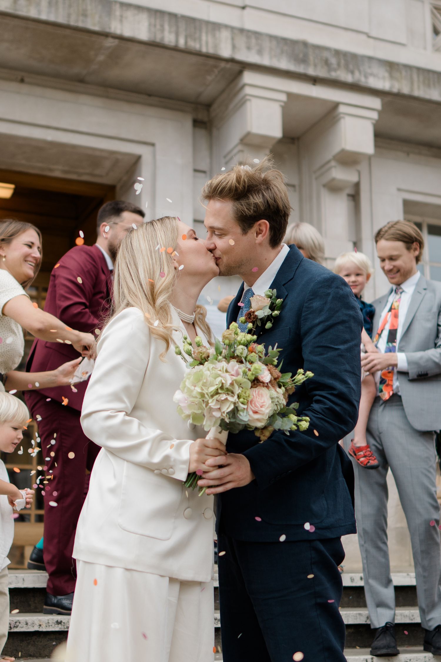 Wedding couple sharing kiss on steps while guests throw confetti in celebration.