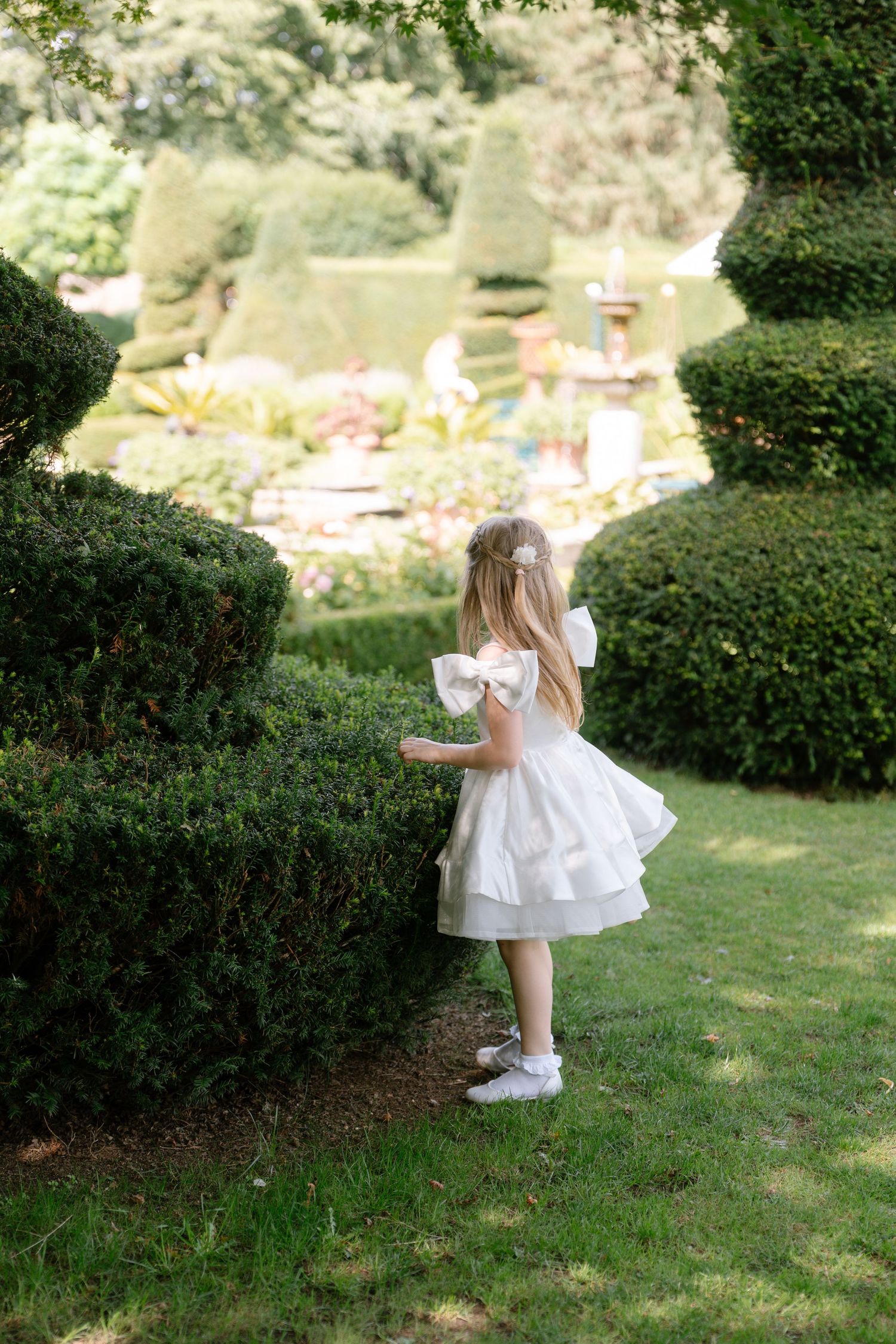 Young girl in a white ruffled dress explores a manicured garden path.
