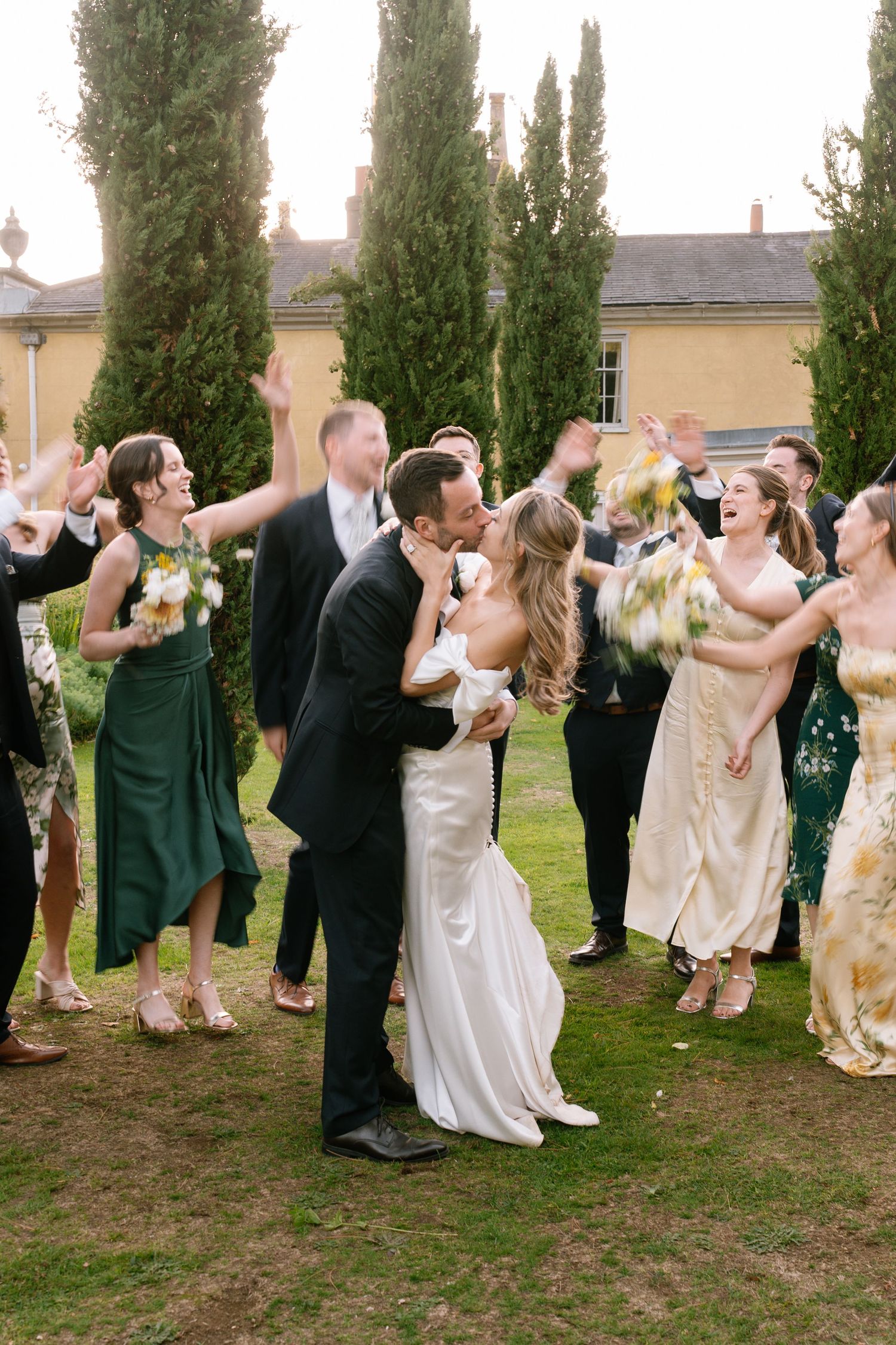 Wedding party performs synchronized celebratory toast outdoors near cypress trees.