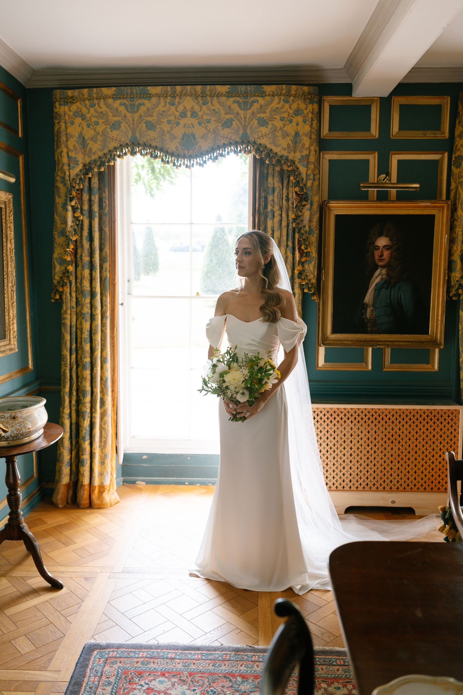 Bride in white gown poses by window with elegant gold and green decor.