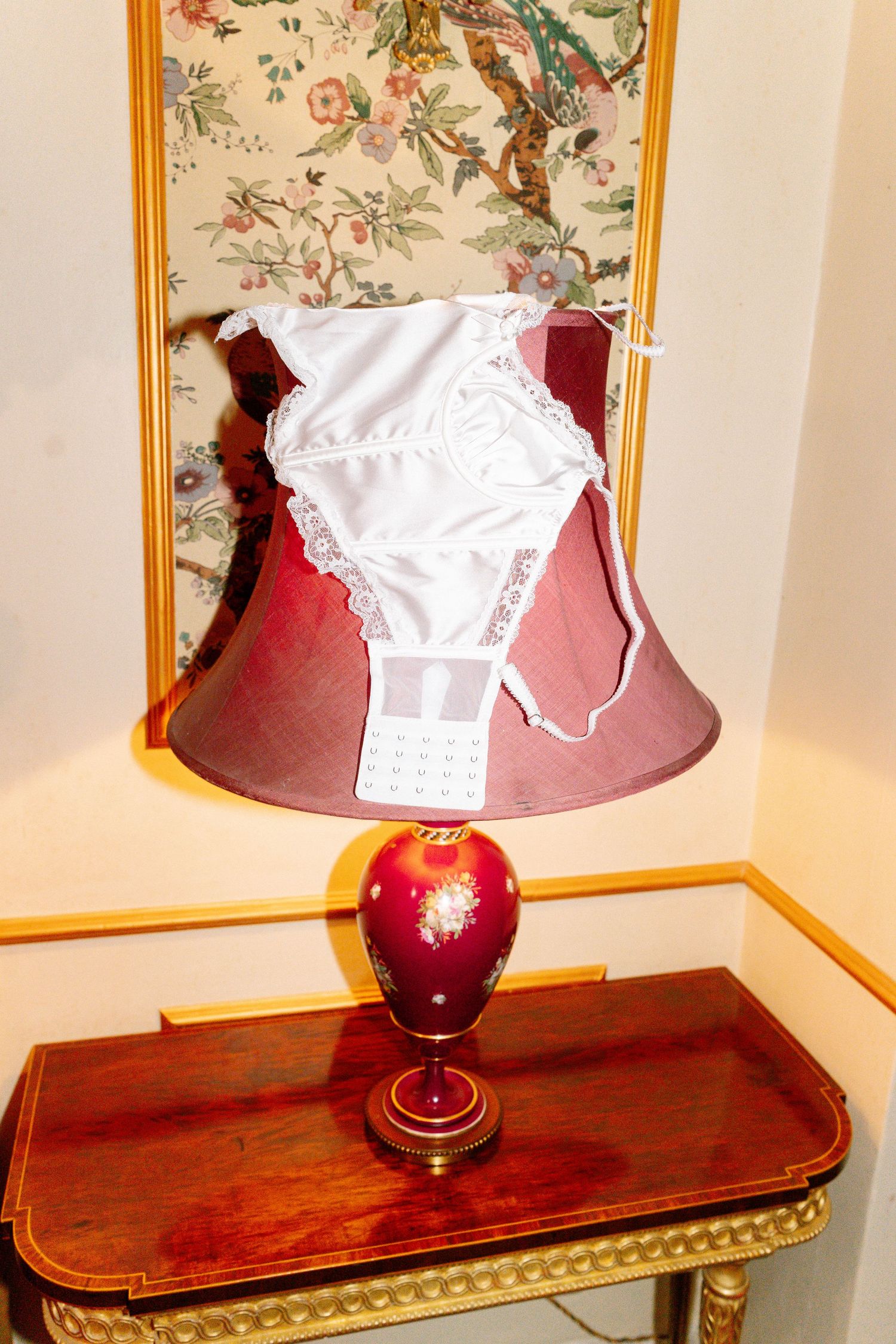 White lace garment displayed on wooden console table with red lamp against floral wallpaper.