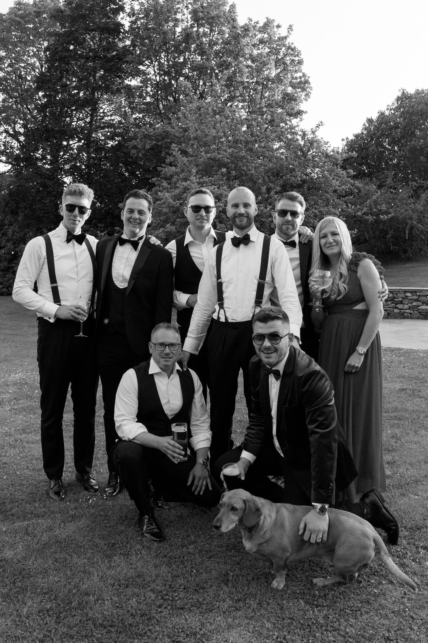 Wedding party in formal attire poses with dog on grass for group photo.