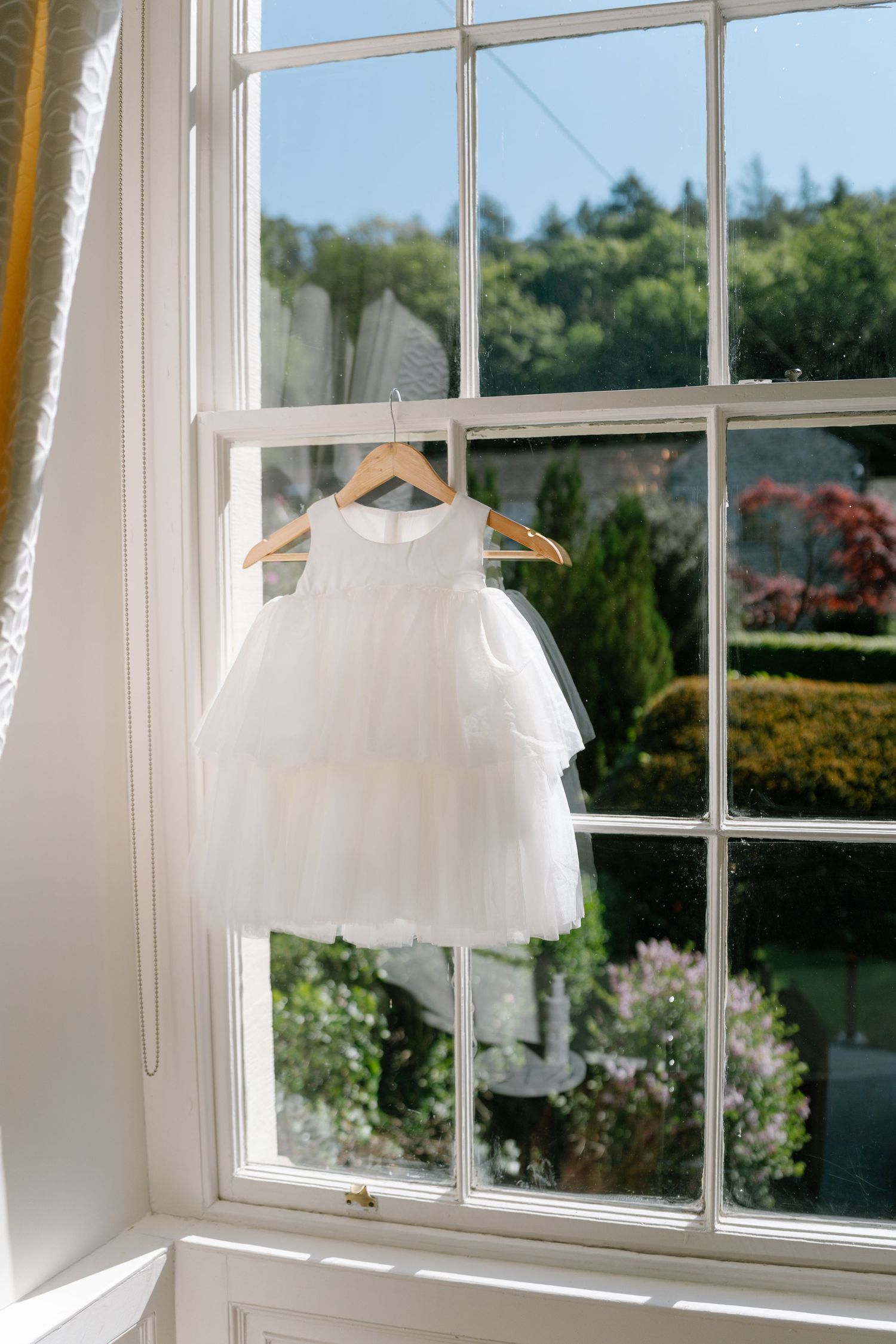 White tulle flower girl dress hanging in sunny window with garden view.