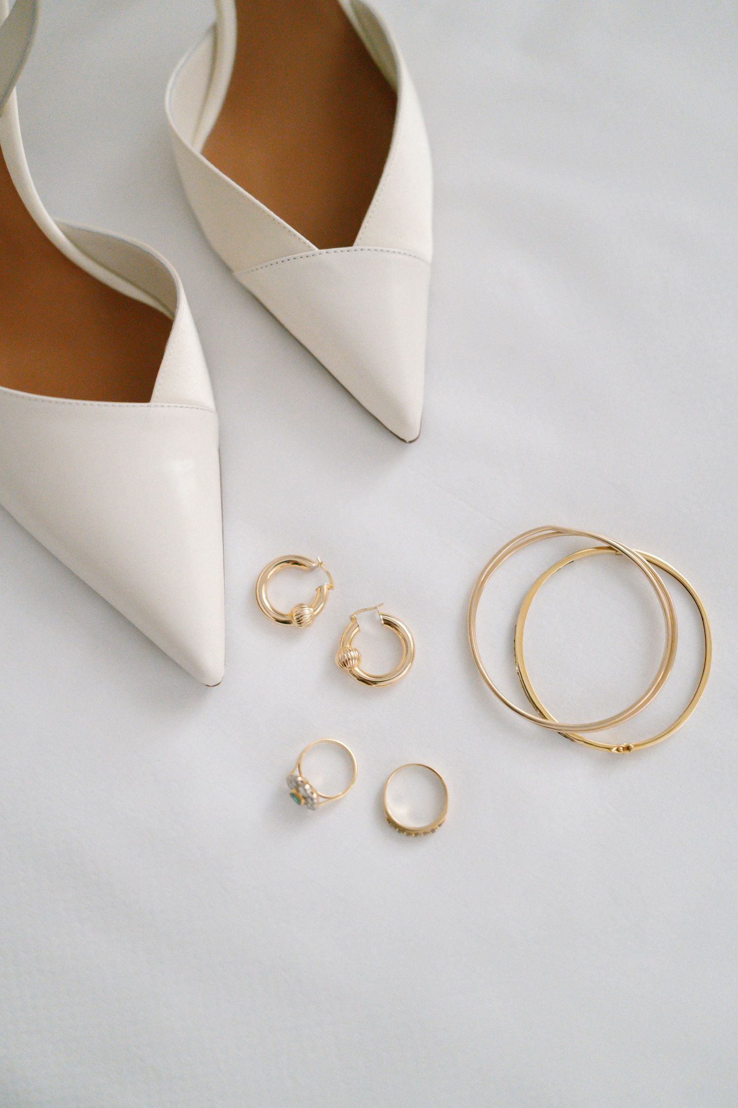 Elegant white bridal shoes with gold jewelry and rings arranged on white surface.