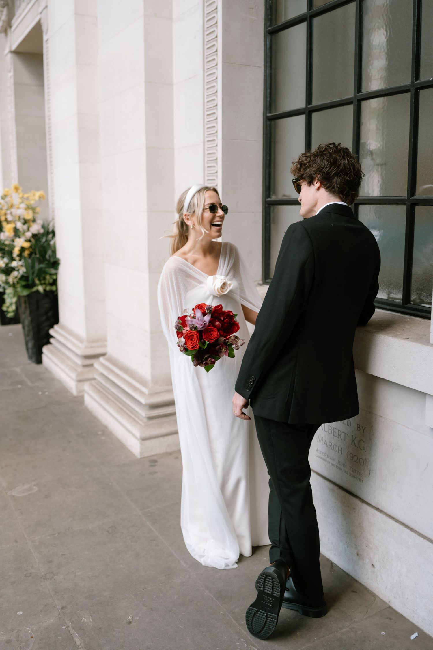 Wedding couple in white dress and black suit playfully interacting near large windows of a classical building.