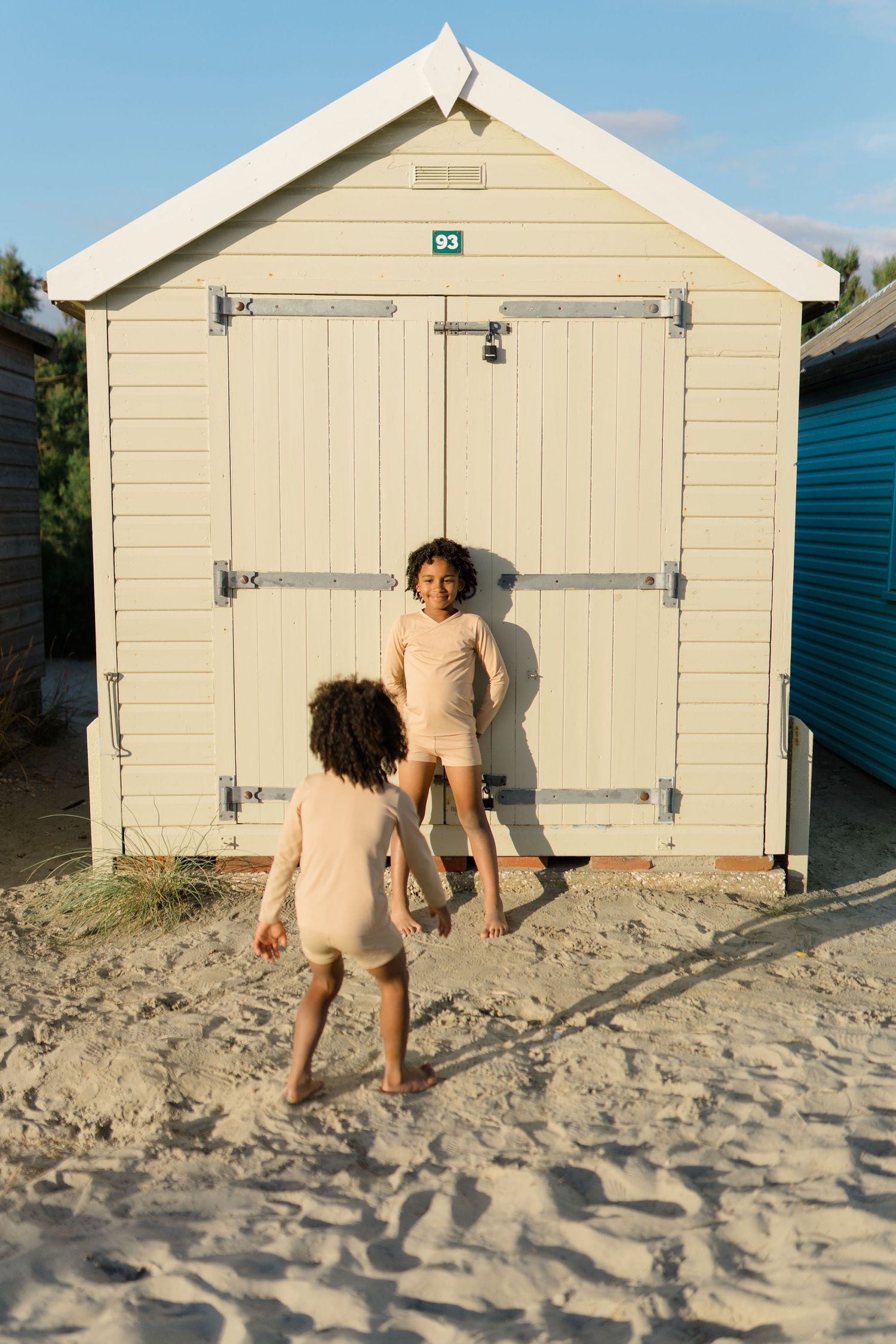 Two figures in beige outfits play in front of a cream-colored beach hut on sandy shores.