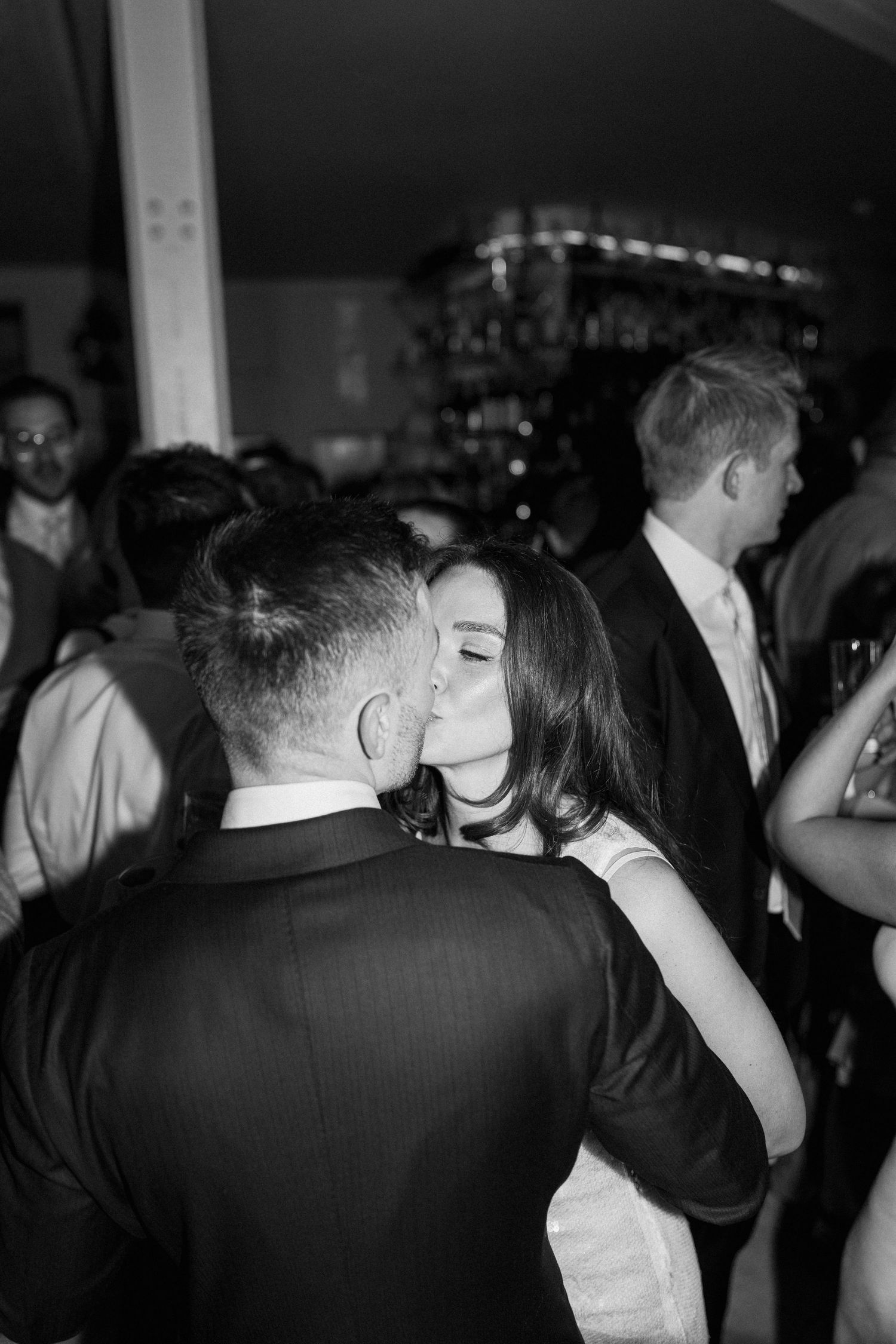Bride and groom share romantic kiss on dance floor surrounded by guests at wedding reception.