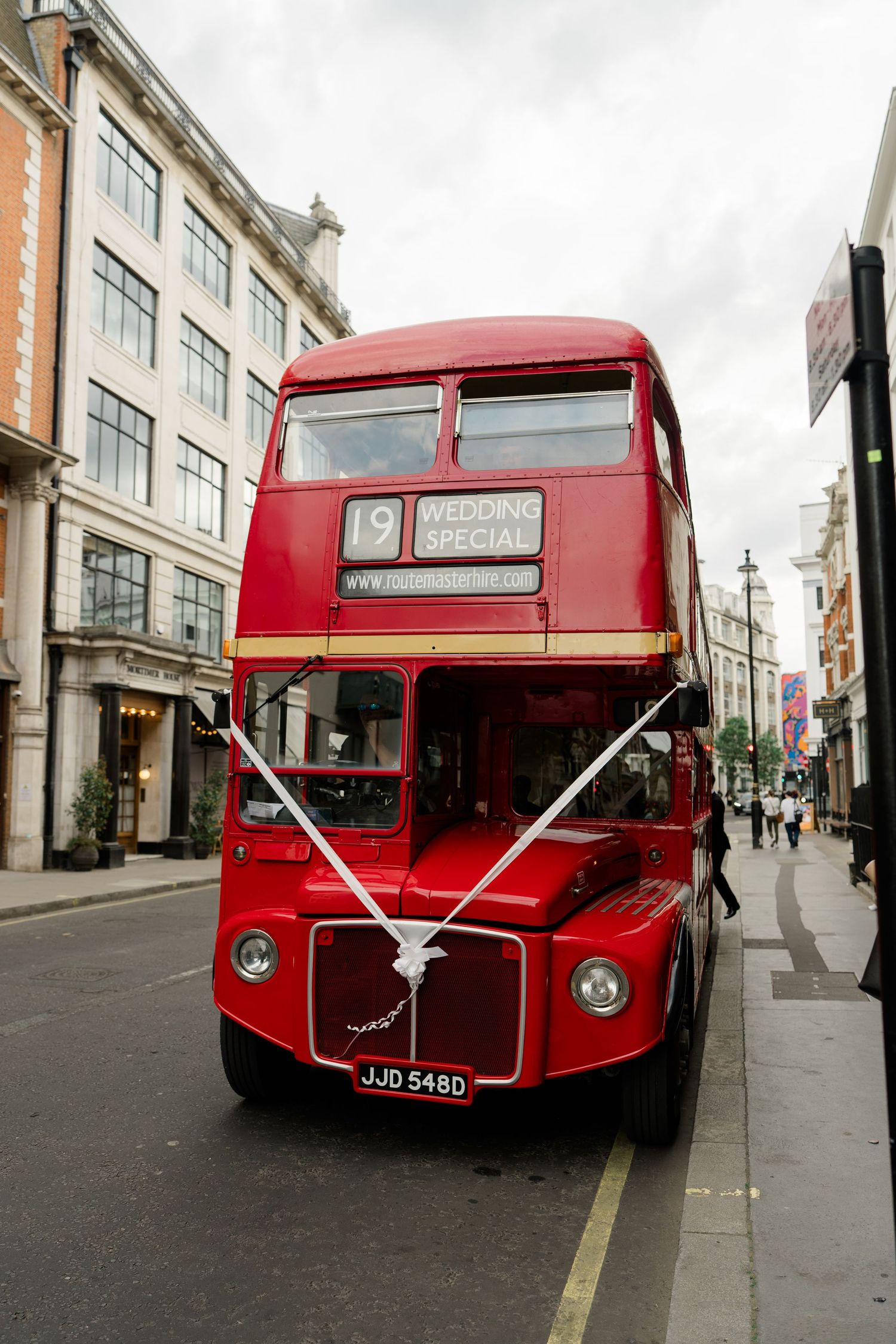 A vintage red London double-decker bus decorated with white ribbons for a wedding special on a city street.