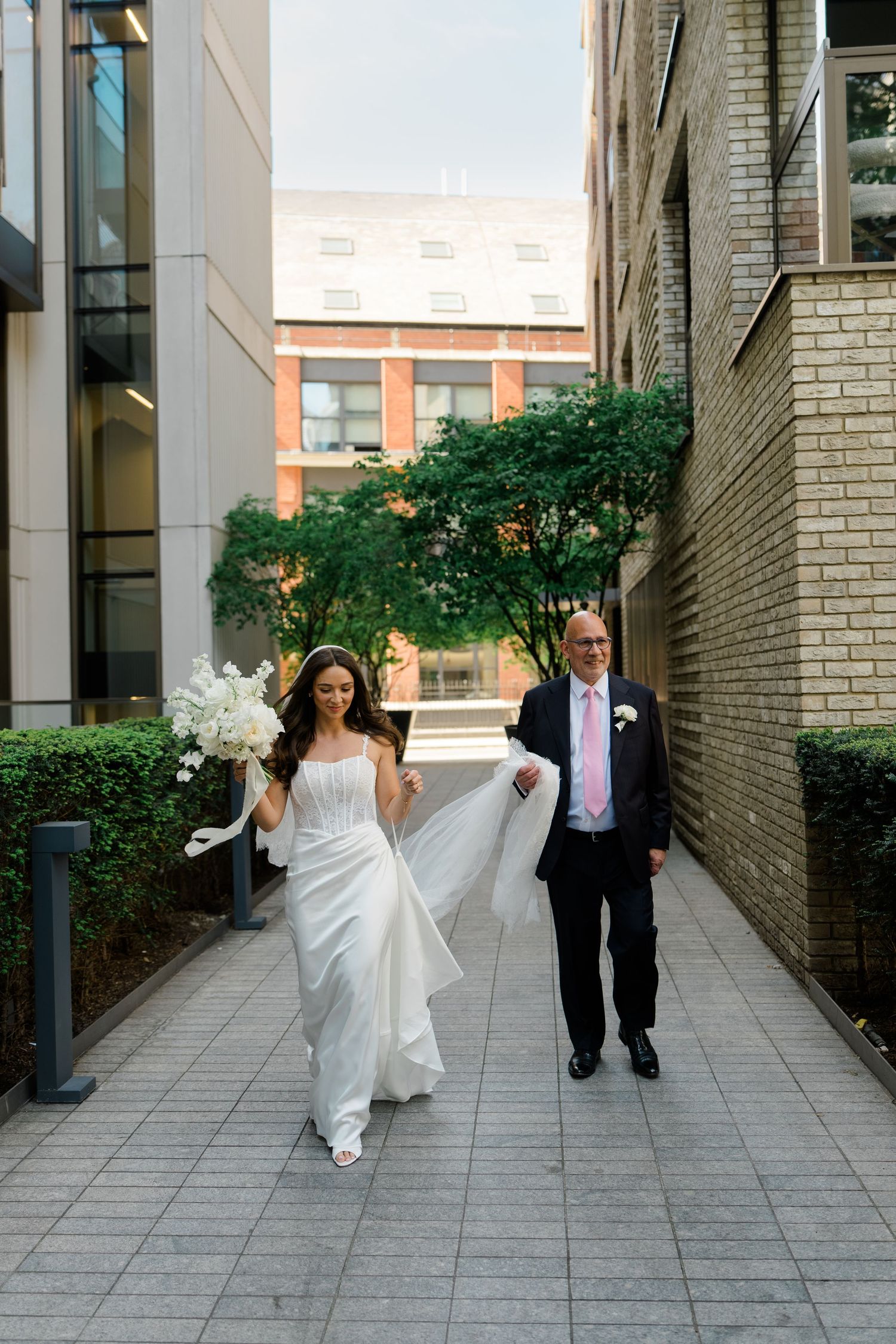 Bride in white gown with veil walks arm-in-arm with her father along an urban pathway.