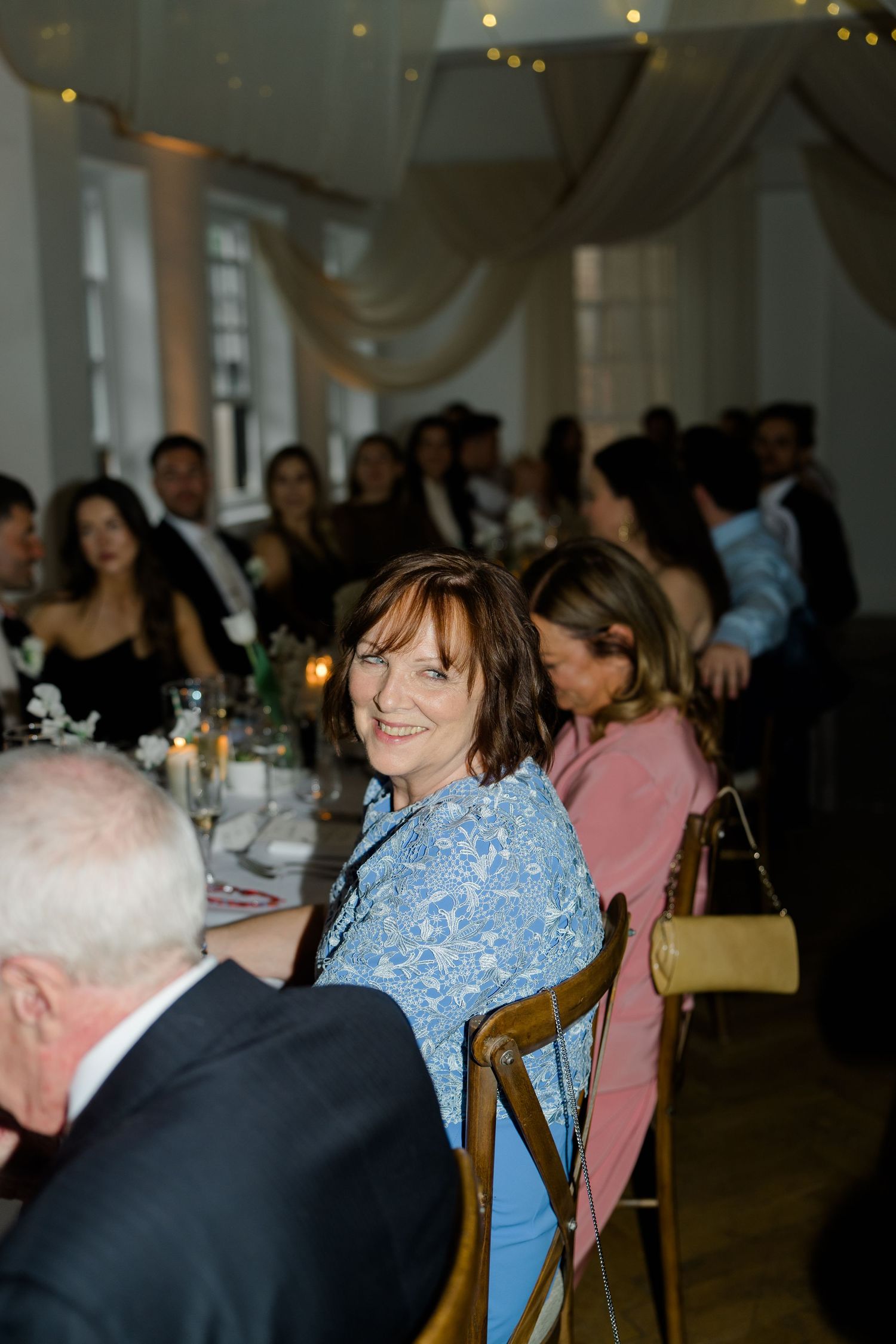 Smiling female wedding guest in a blue floral dress seated at a candlelit wedding reception dinner table.