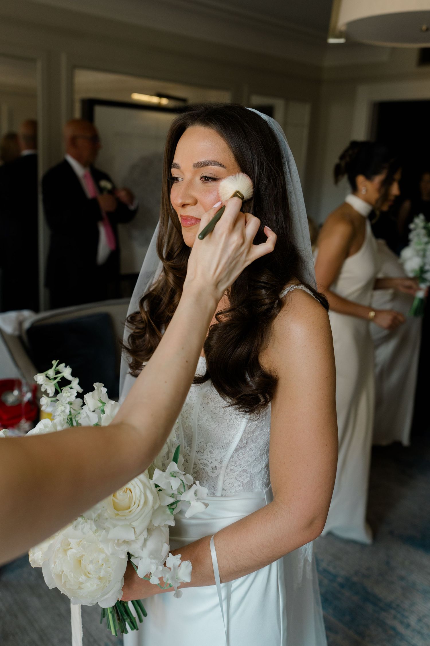 A smiling bride in a white gown and veil receives final makeup touch-ups while holding a bouquet.