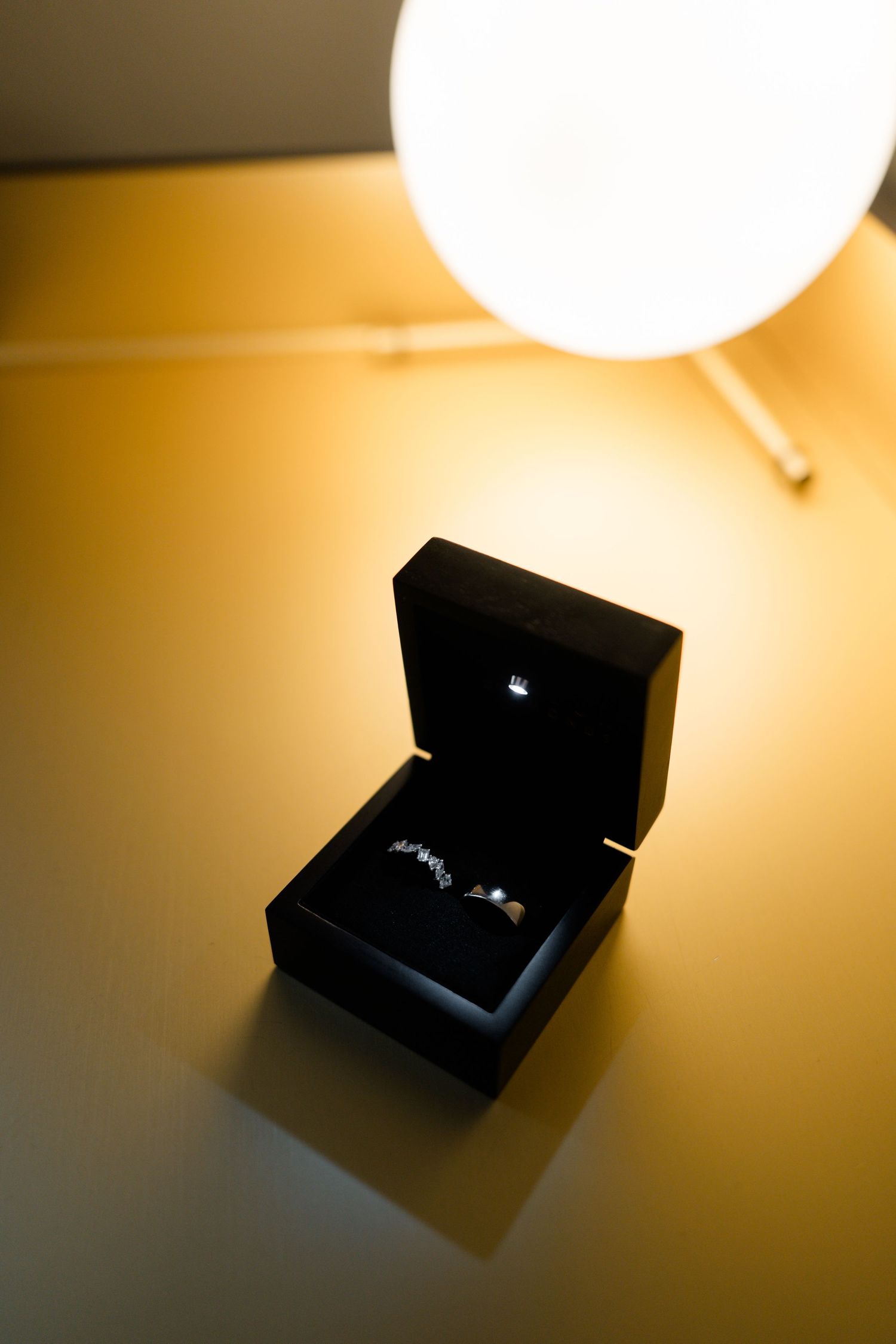 Diamond wedding ring in open black jewelry box illuminated by warm golden lamp light on reflective surface.