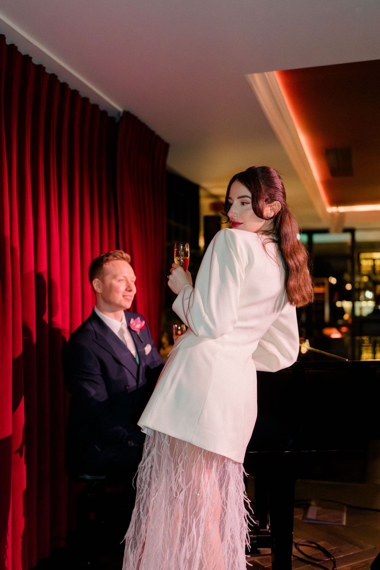 Bride in white feathered dress holds champagne flute at piano bar while groom watches admiringly.