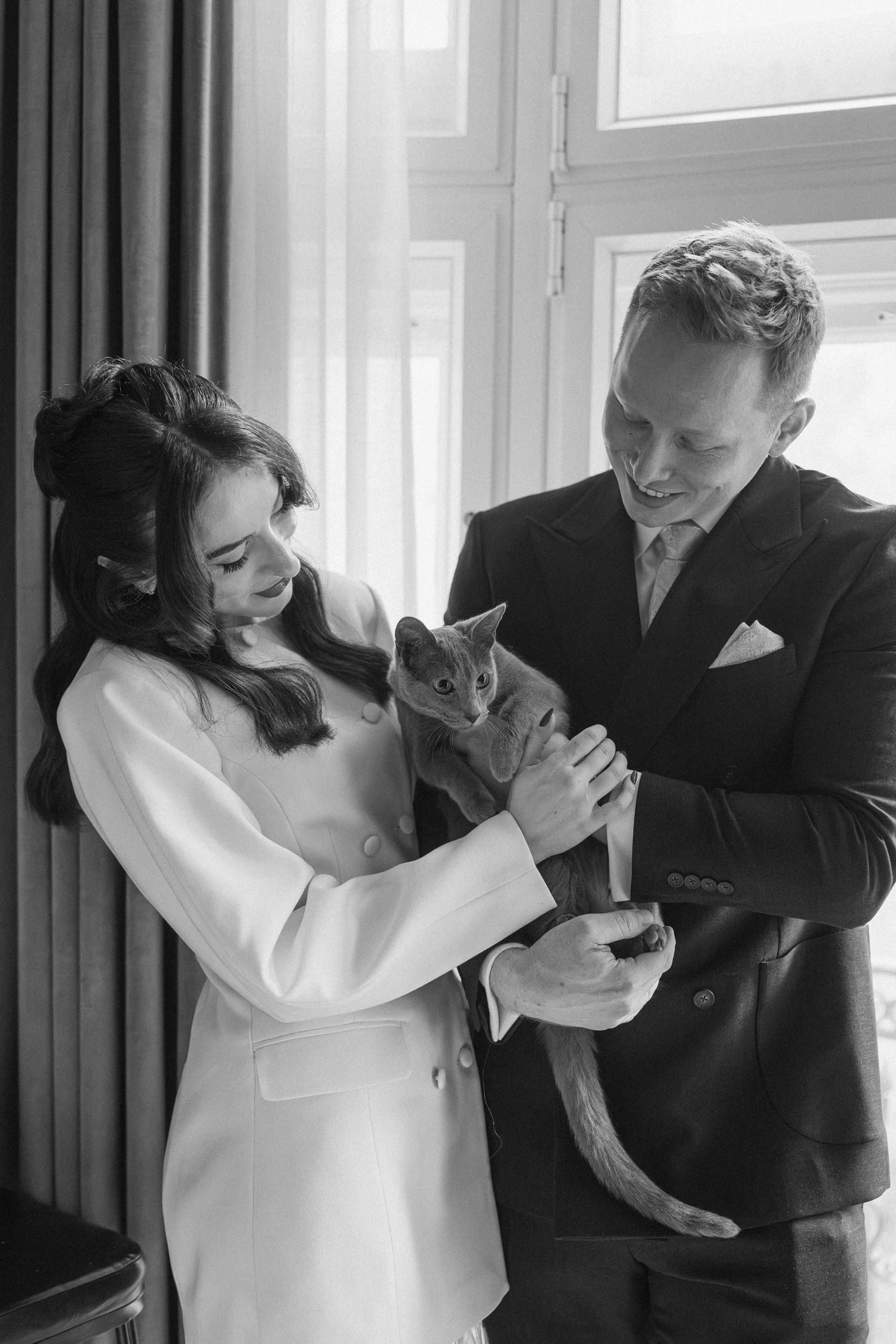 Black and white photo of a bride and groom holding a cat near a window on their wedding day.