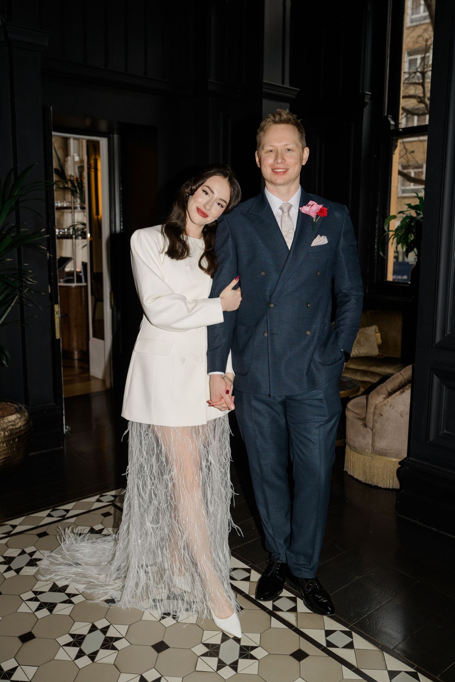 Bride in white feather mini dress and groom in navy suit posing on black and white tile floor.