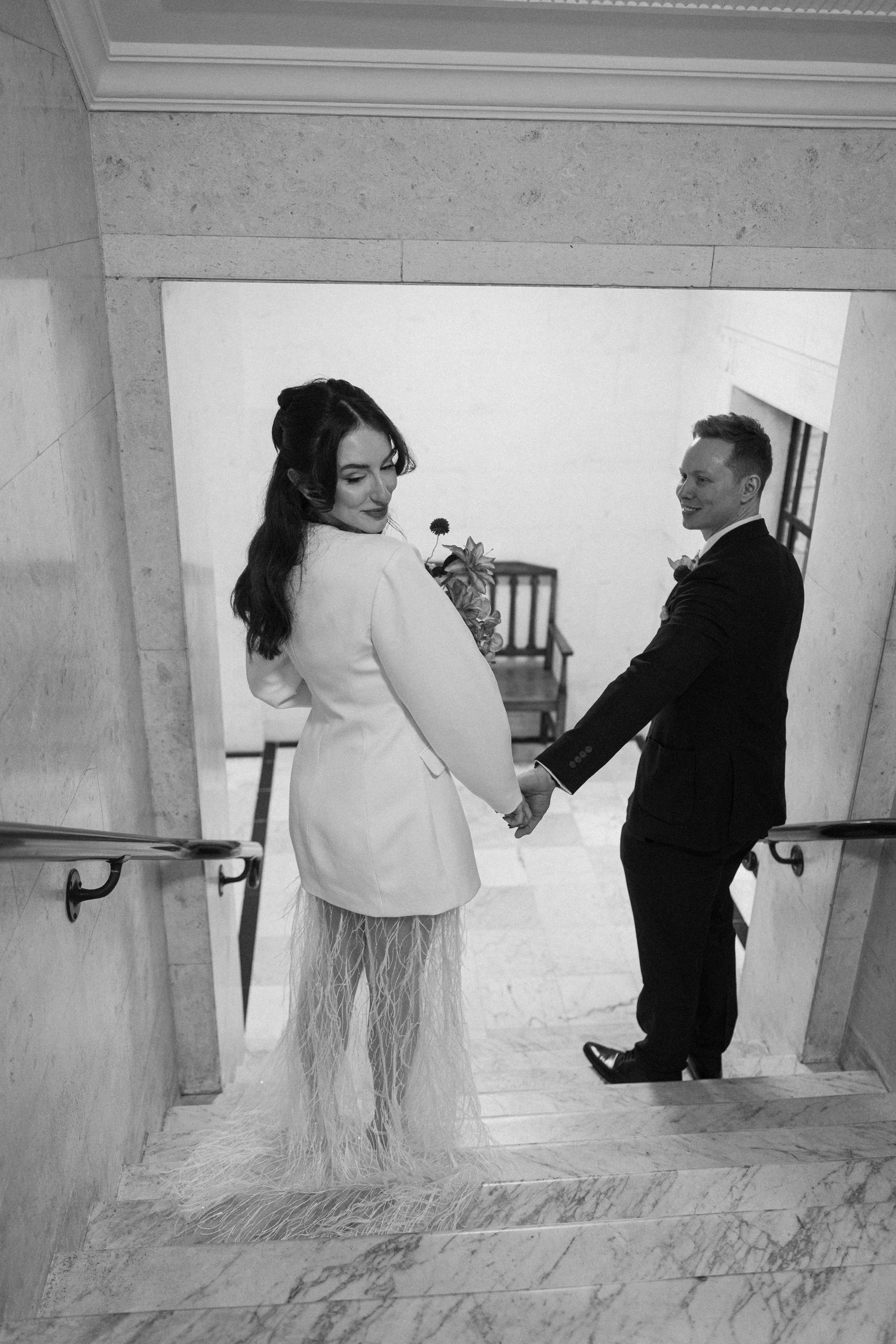 Black and white photo of bride in white blazer and feather skirt holding hands with groom on marble staircase.