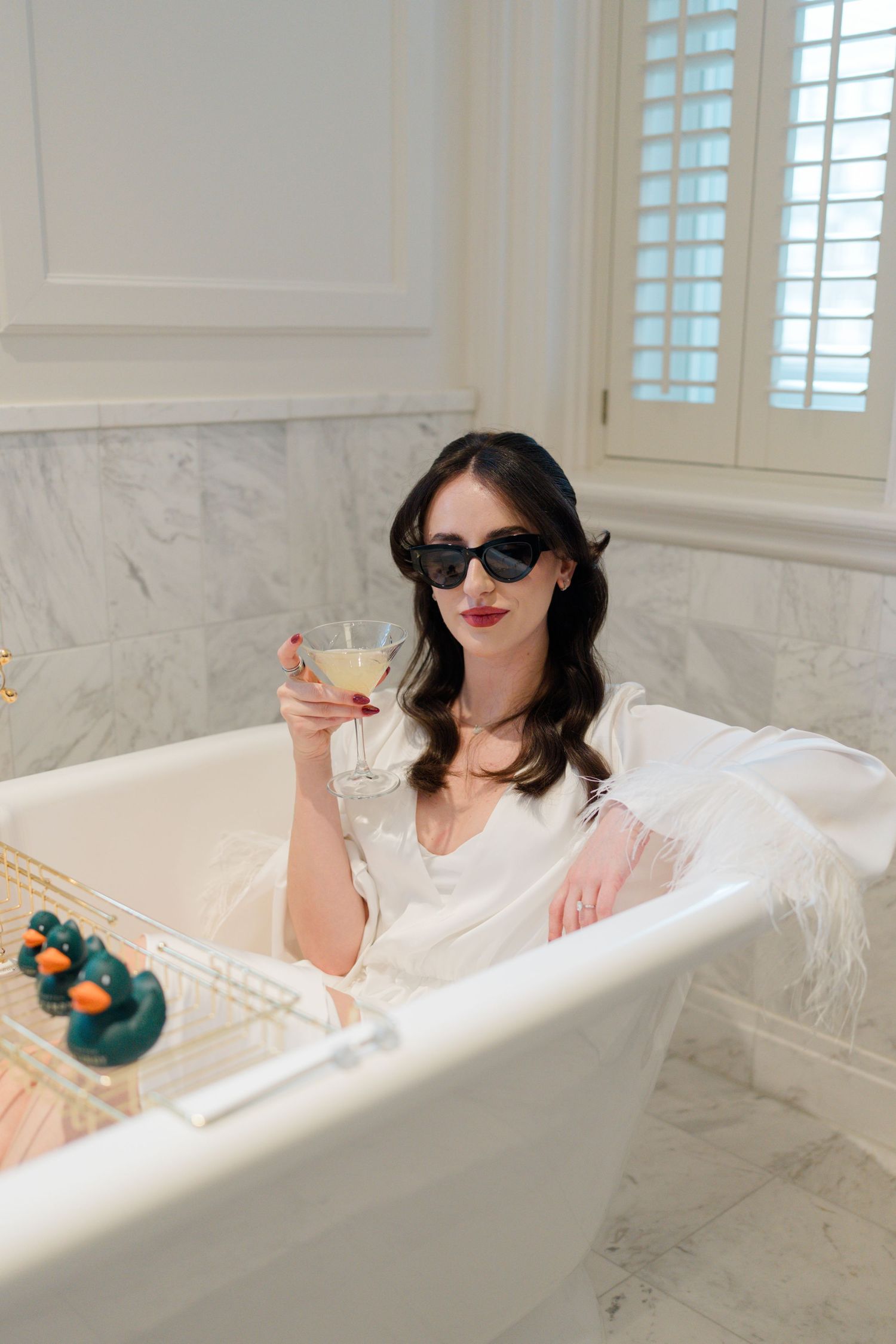 Bride in white robe and sunglasses relaxing in marble bathtub holding champagne glass.