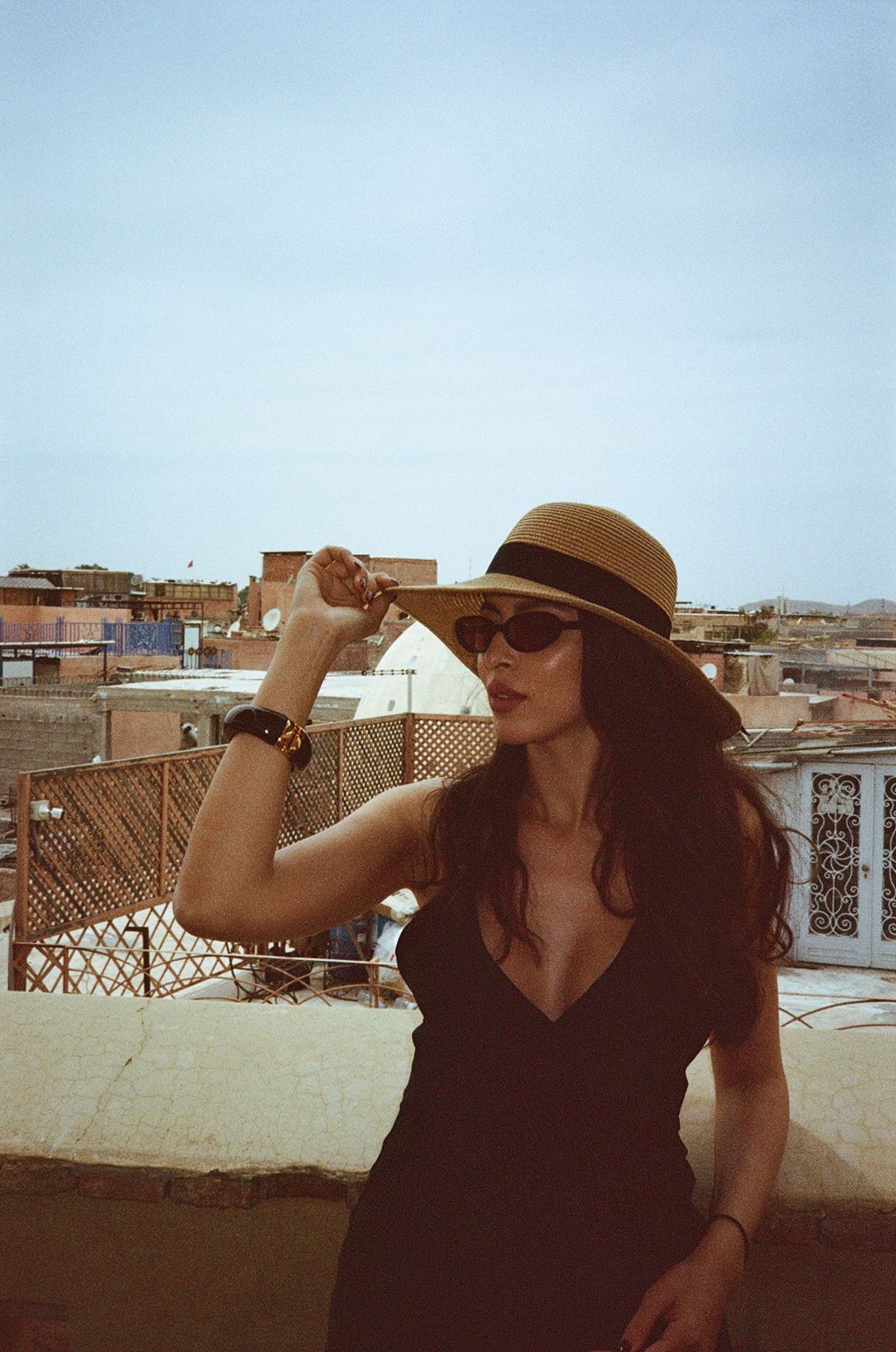 Woman in black dress and straw hat poses on rooftop terrace with sunglasses, city skyline in background.