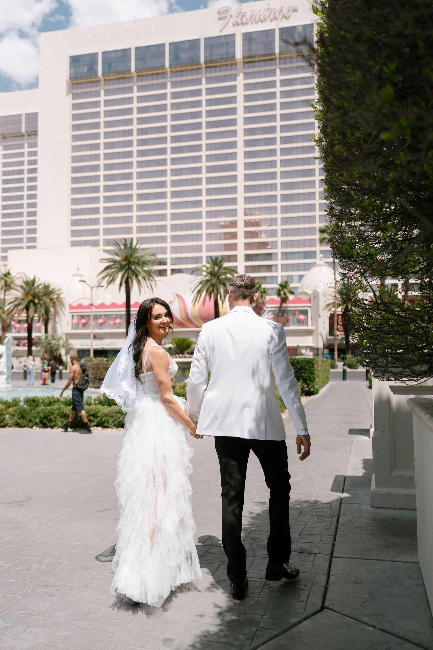A couple in wedding attire walking away from the Flamingo hotel with palm trees in view.