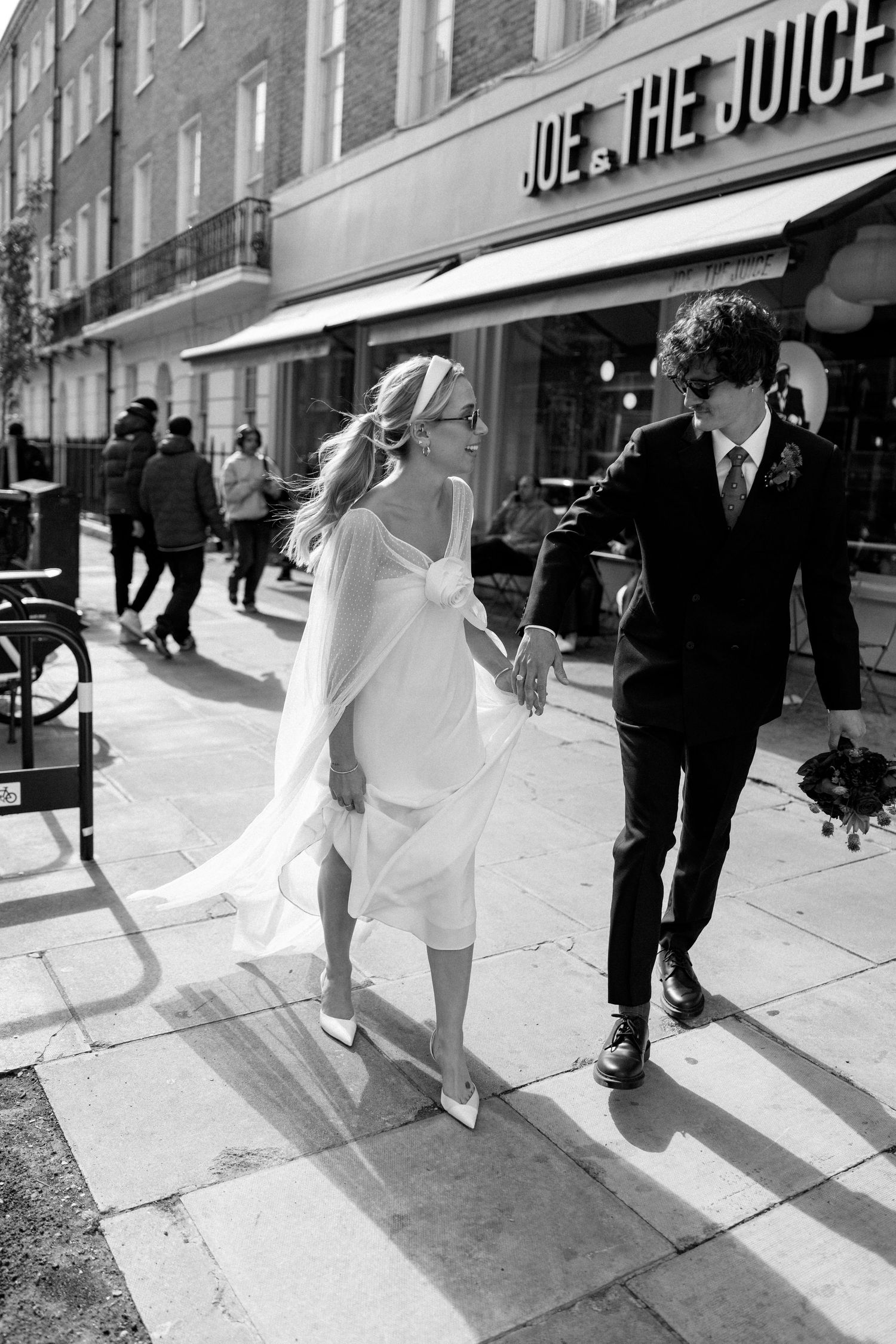 A couple in wedding attire walks past Joe & The Juice cafe on a city street.
