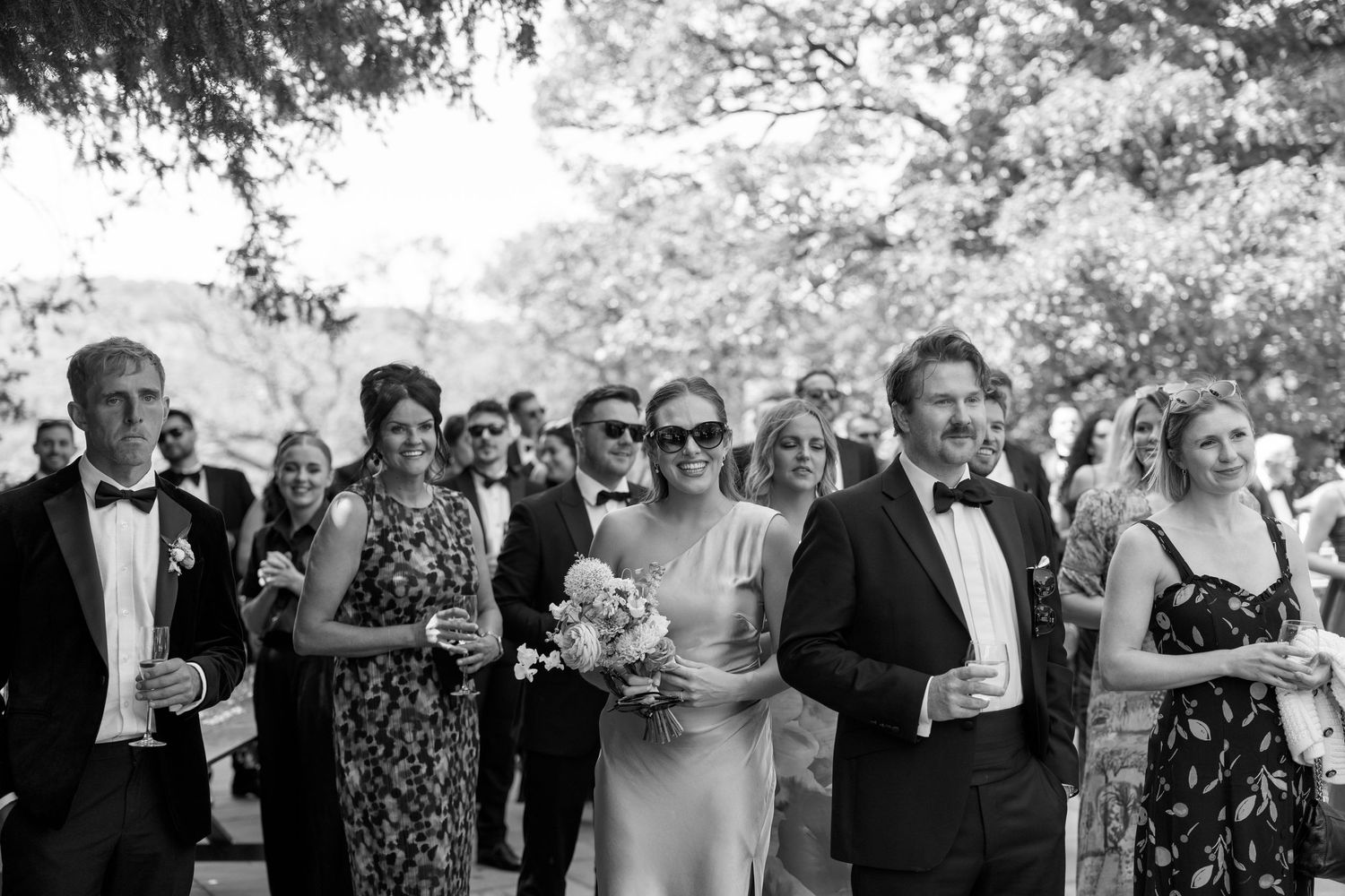 Wedding guests in formal attire watch the celebration proceedings in black and white.