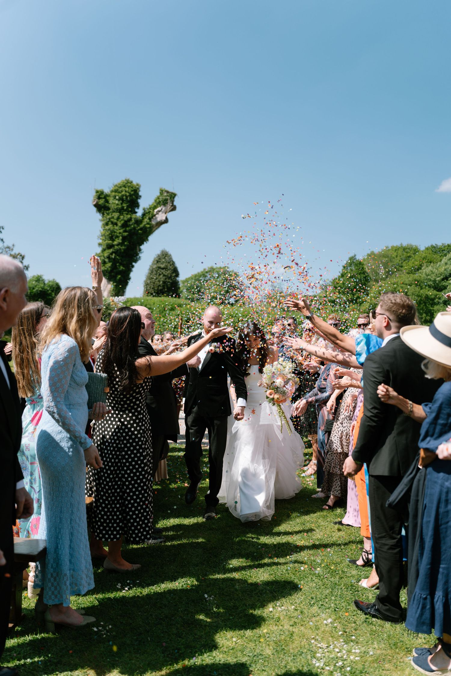Wedding guests toss colorful confetti in celebration under a bright blue sky.