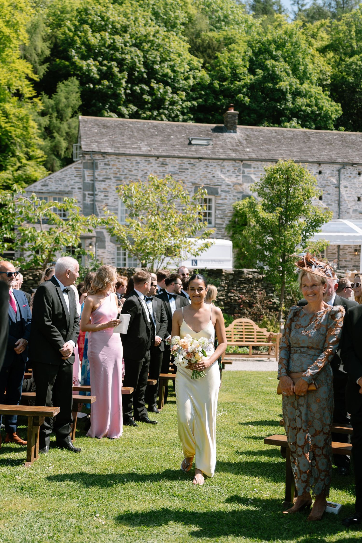 Wedding ceremony guests gather on a lawn in front of stone buildings on a sunny day.