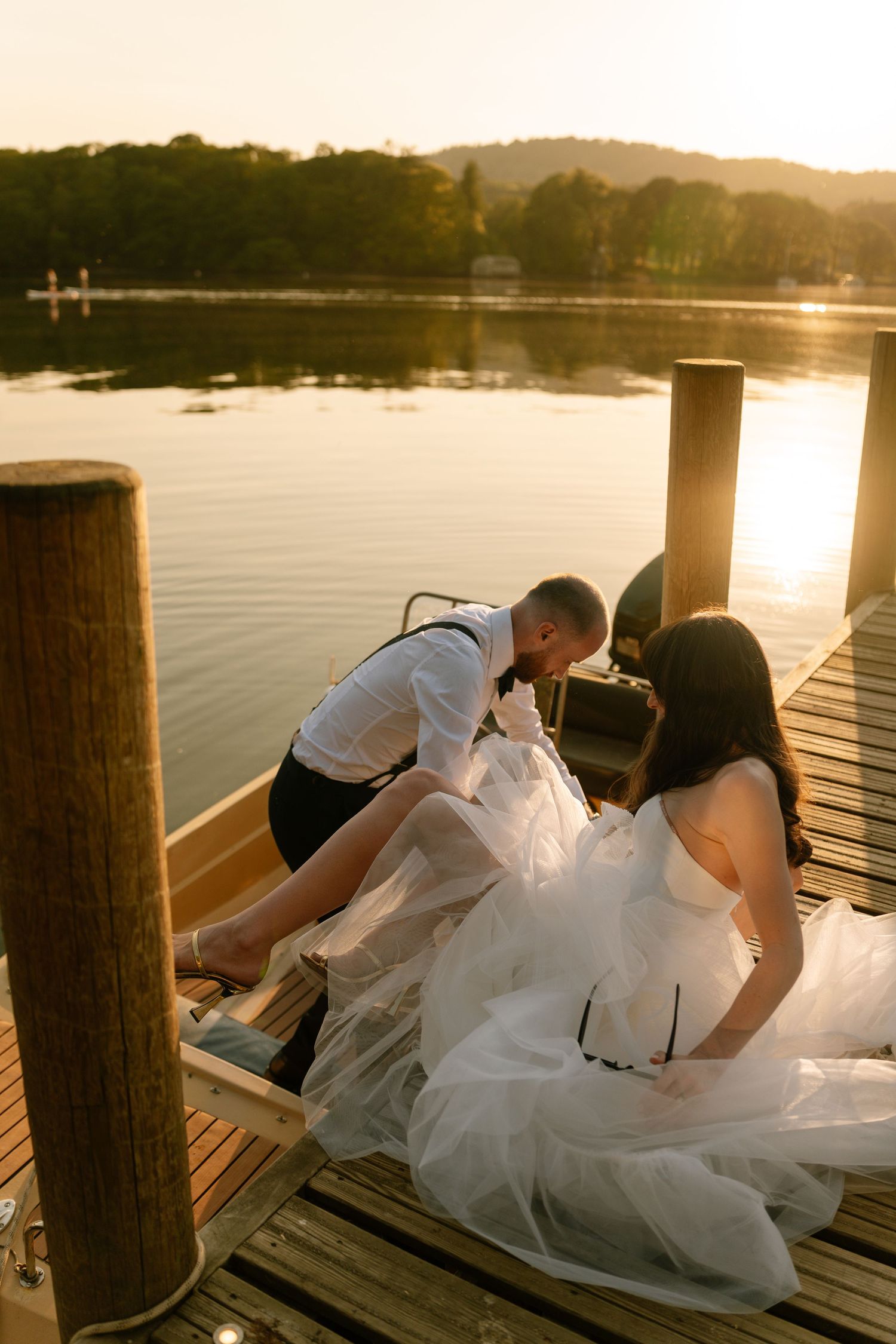 Couple in wedding attire reclining on wooden dock during sunset photoshoot.