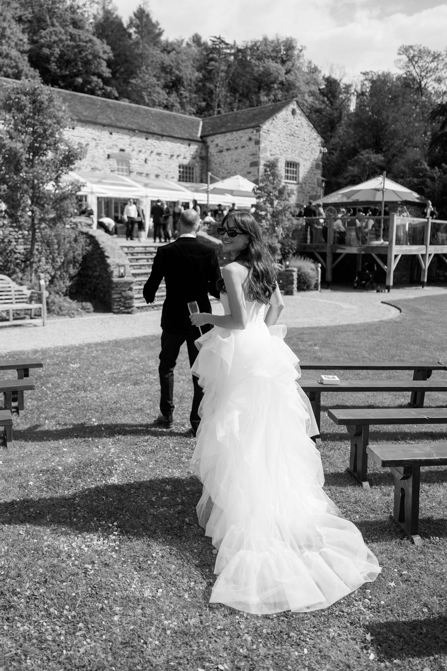 A bride in a flowing white gown walks across a grassy area with wooden benches.