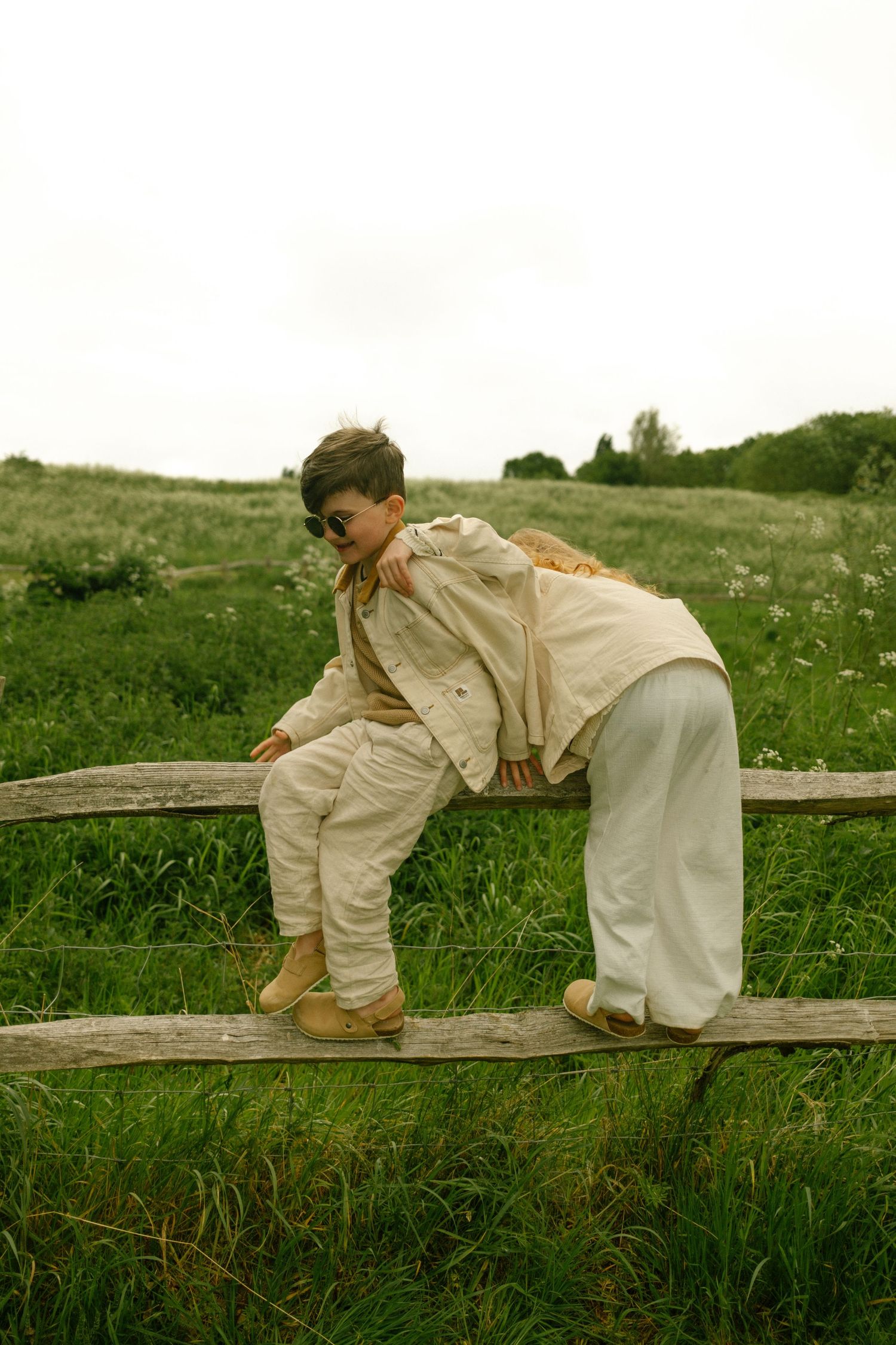Two people in cream-colored outfits climb and play on wooden fence posts in a meadow.