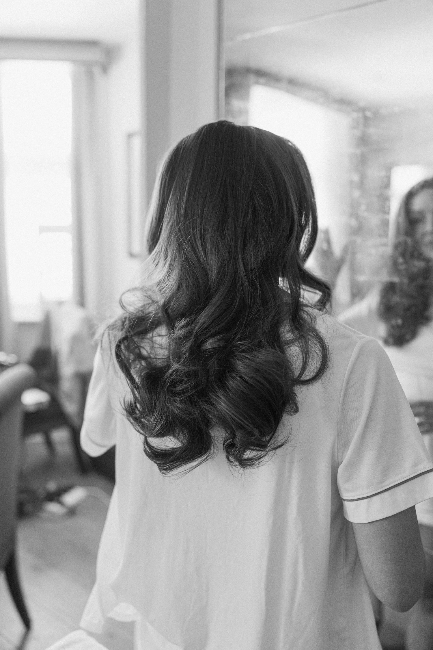 Black and white photo of a bride with long curly hair viewed from behind in a salon.