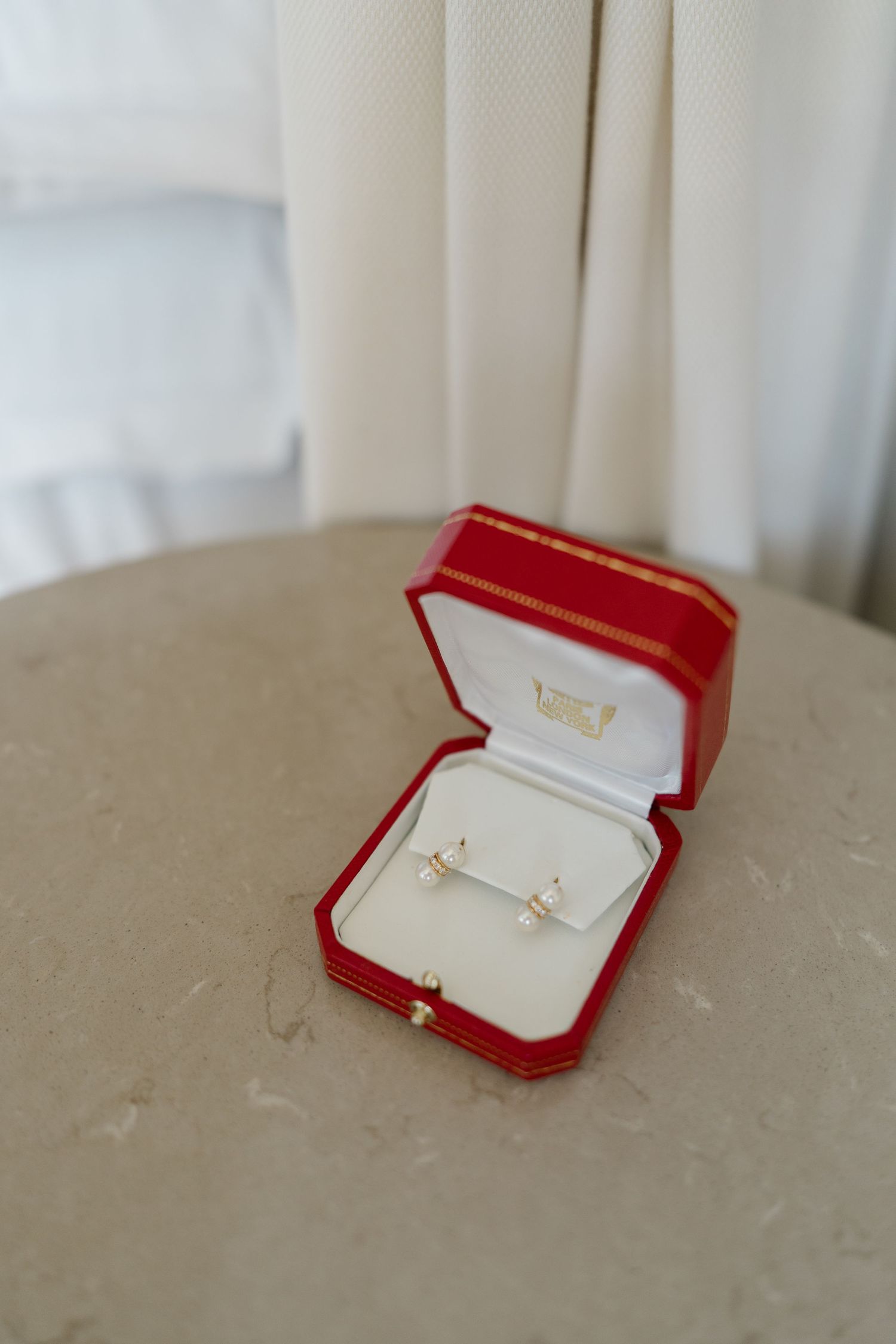 Gold stud earrings with pearl drops displayed in an open red Cartier jewelry box on a marble surface.