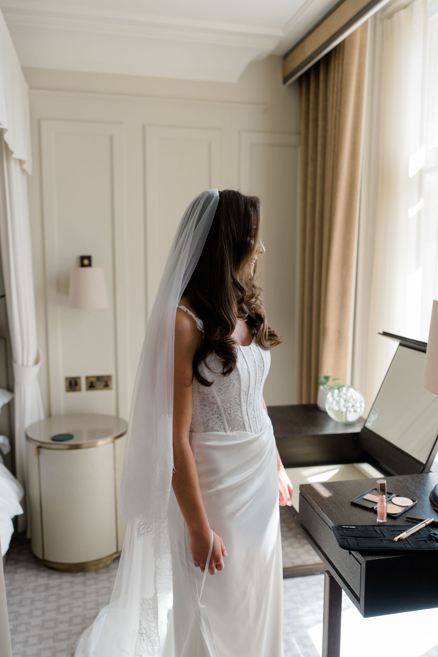 Bride in a white lace wedding gown and veil standing by a vanity mirror in a hotel room.