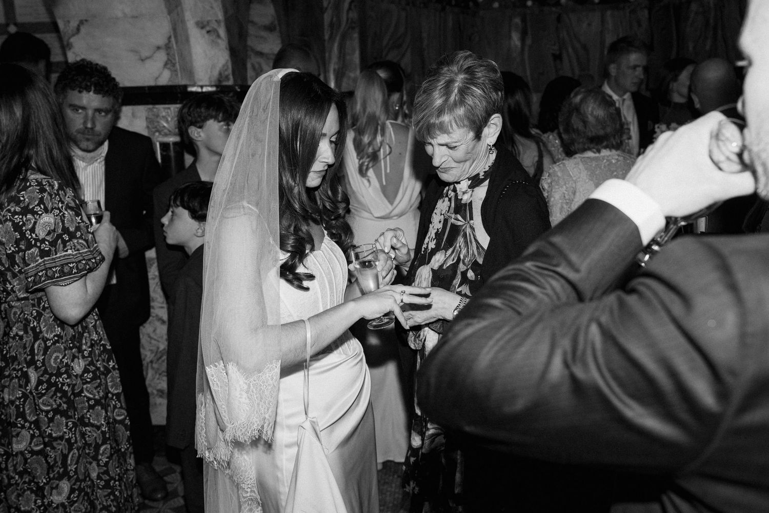 Black and white photo of bride in veil receiving a ring from a guest at her wedding.