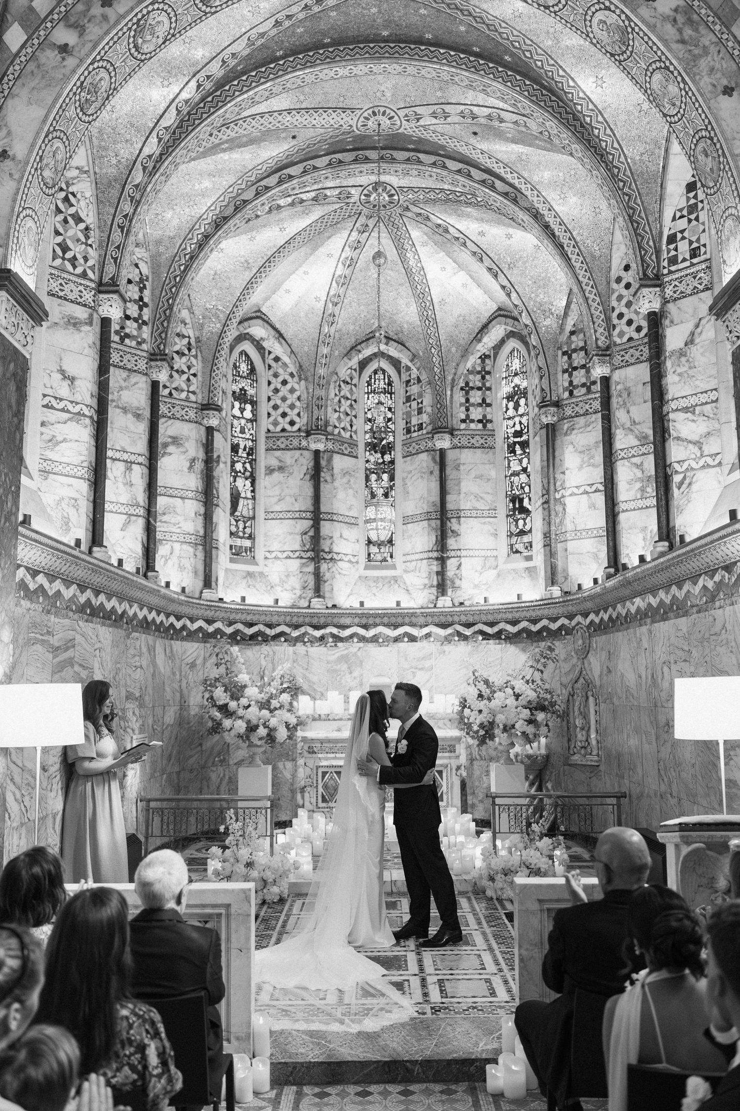 Black and white photo of a bride and groom sharing their first kiss at the altar of an ornate chapel.