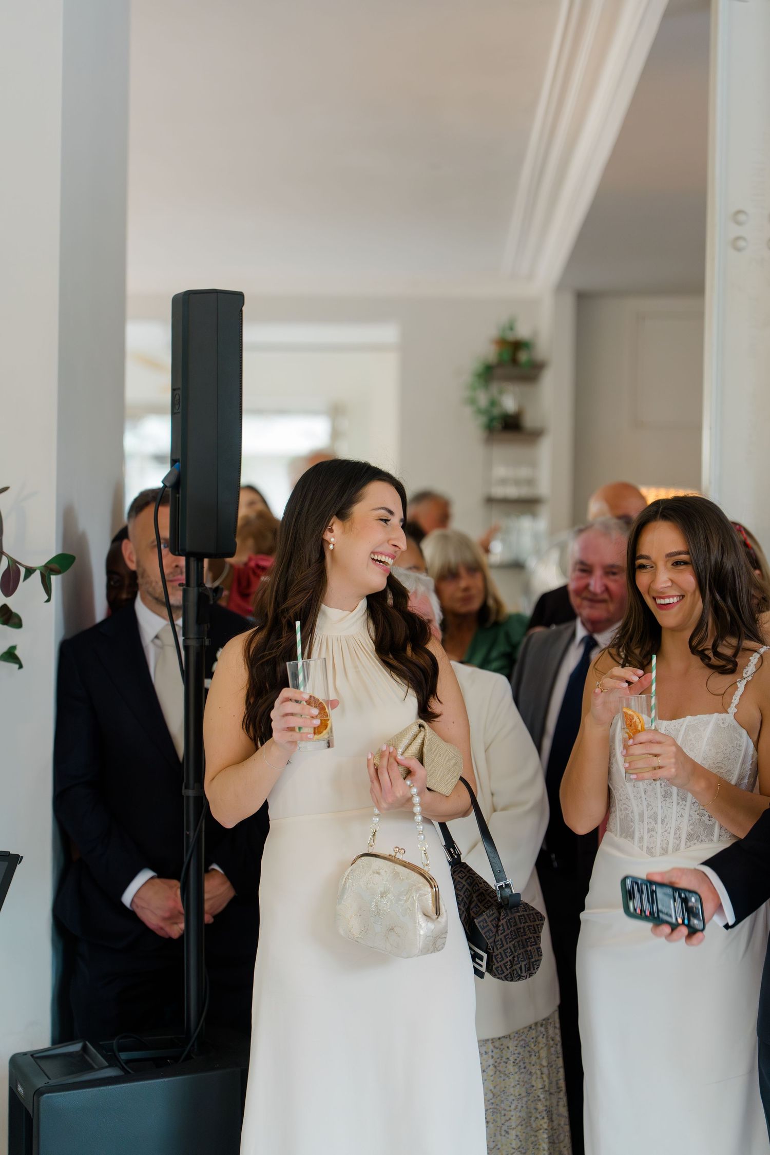Two women in white dresses holding champagne glasses and smiling at a wedding reception.