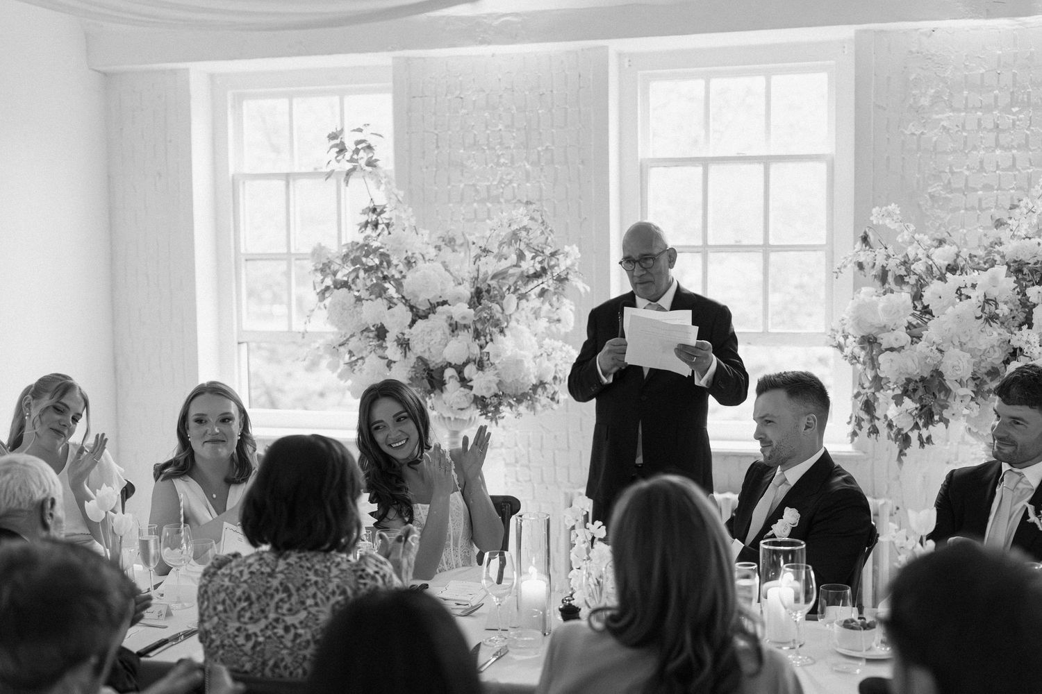 Black and white photo of a man giving a speech at a wedding reception with floral arrangements.