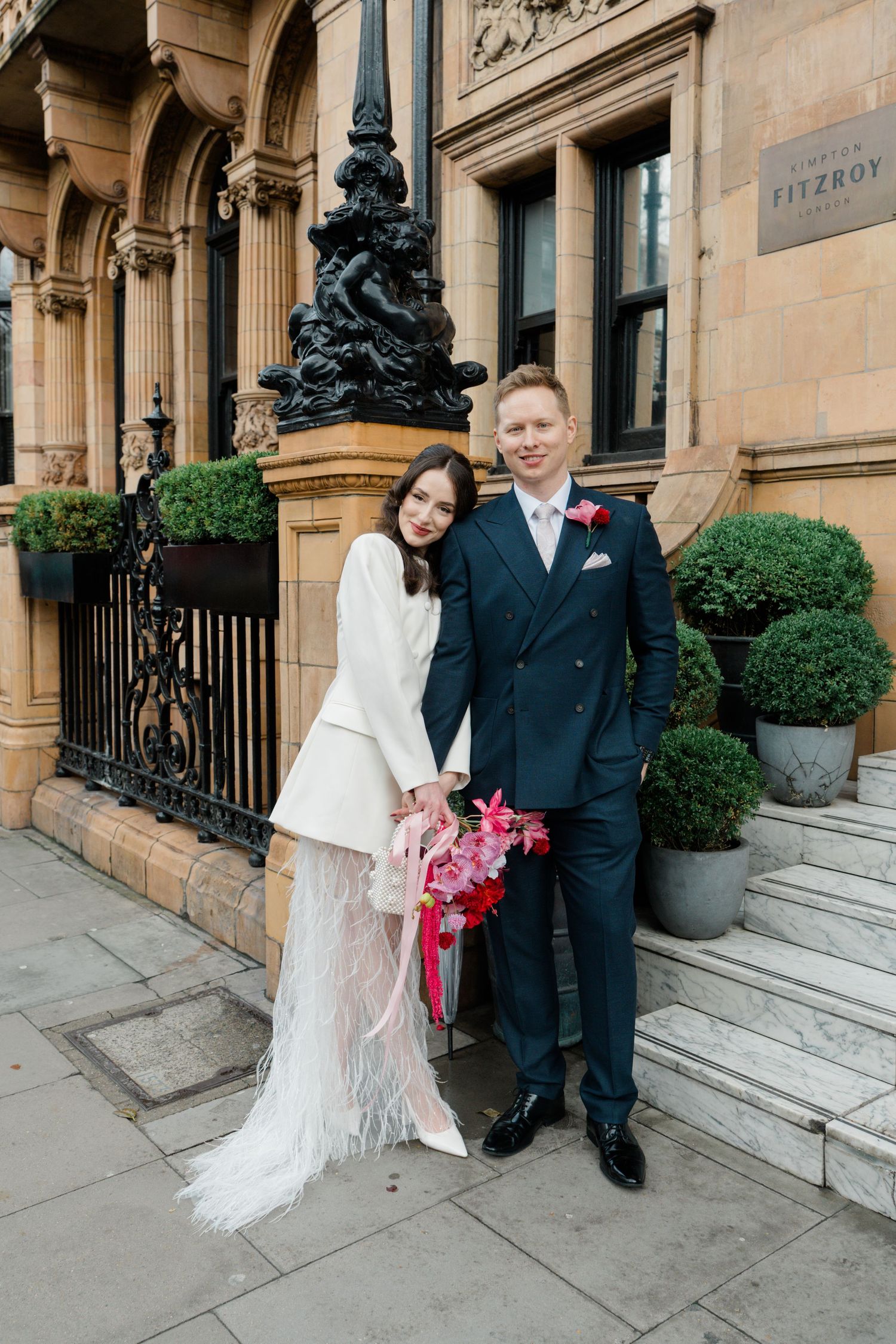 Bride and groom posing outside Hotel Fitzroy in London, bride wearing white feathered skirt suit.