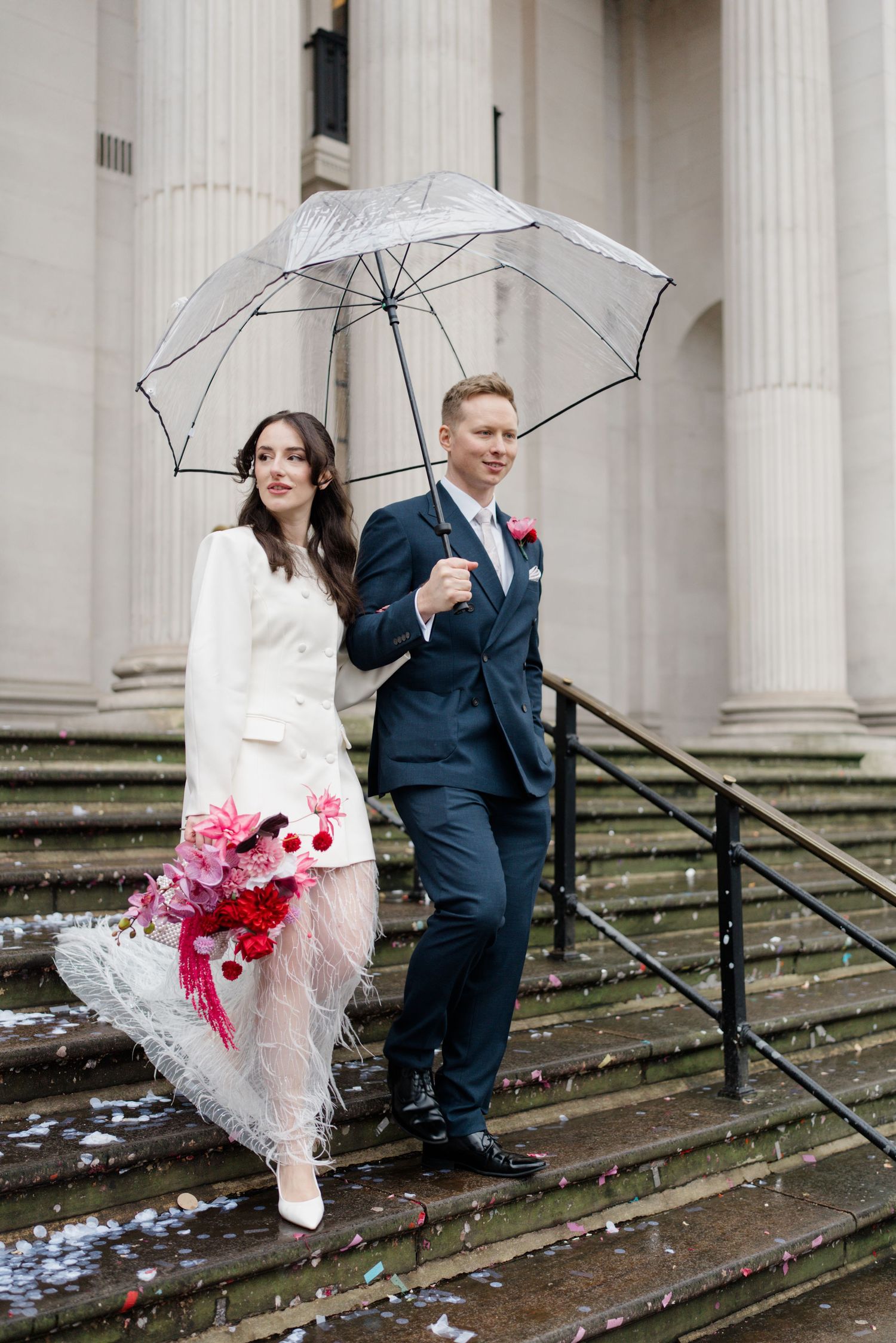 Bride in white feather skirt and groom in navy suit share clear umbrella on rainy wedding day steps.