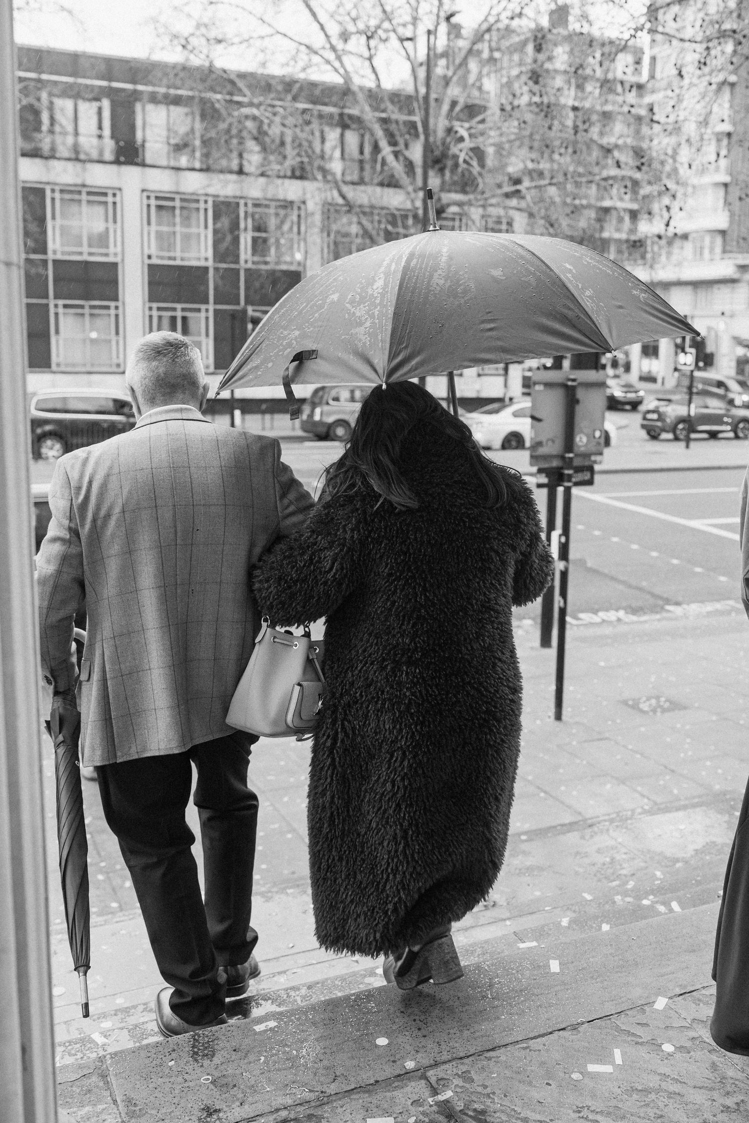 Black and white photo of elderly couple walking under umbrella on a rainy city street.