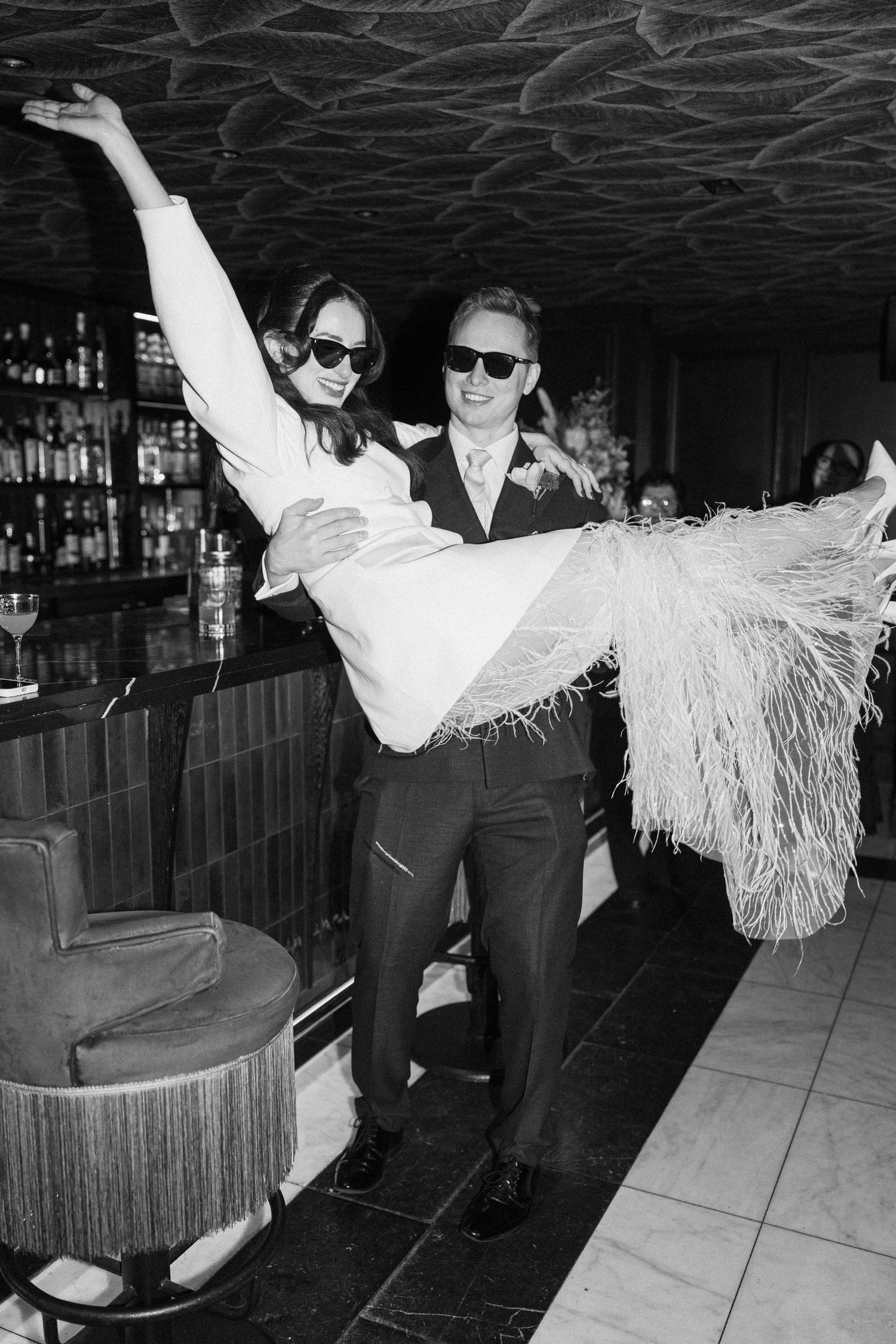 Groom carries bride in feathered dress at bar in black and white wedding photo, both wearing sunglasses.