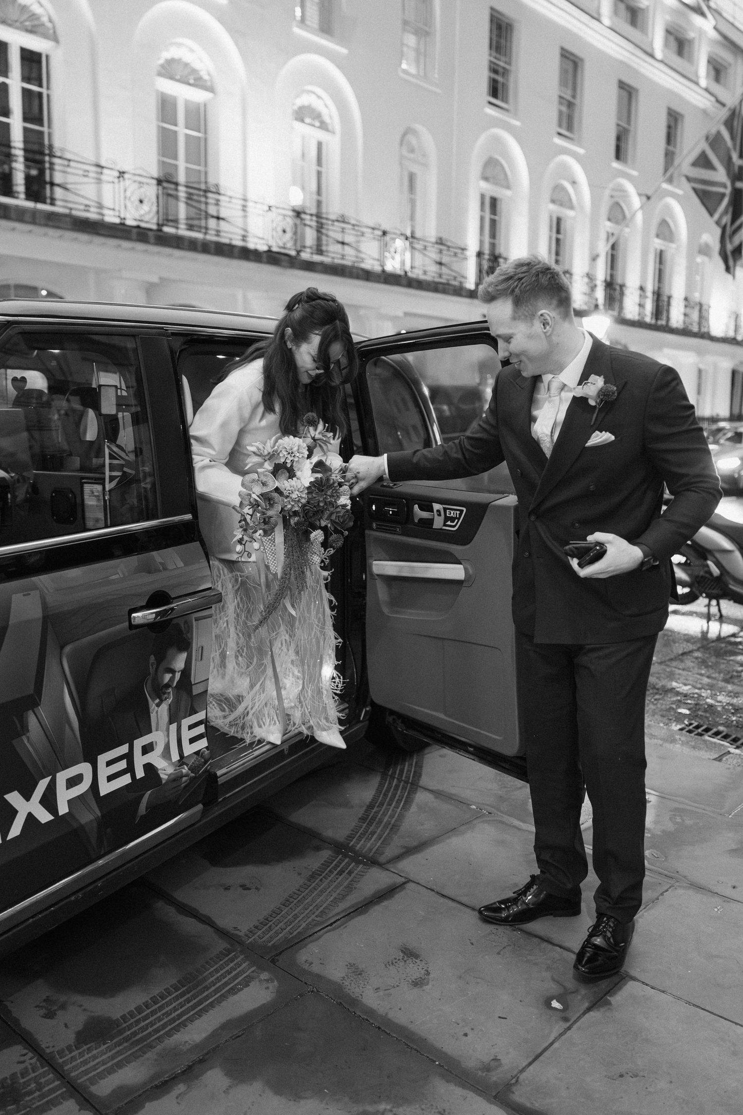 Groom helping bride exit black London taxi cab in black and white wedding photo at night.