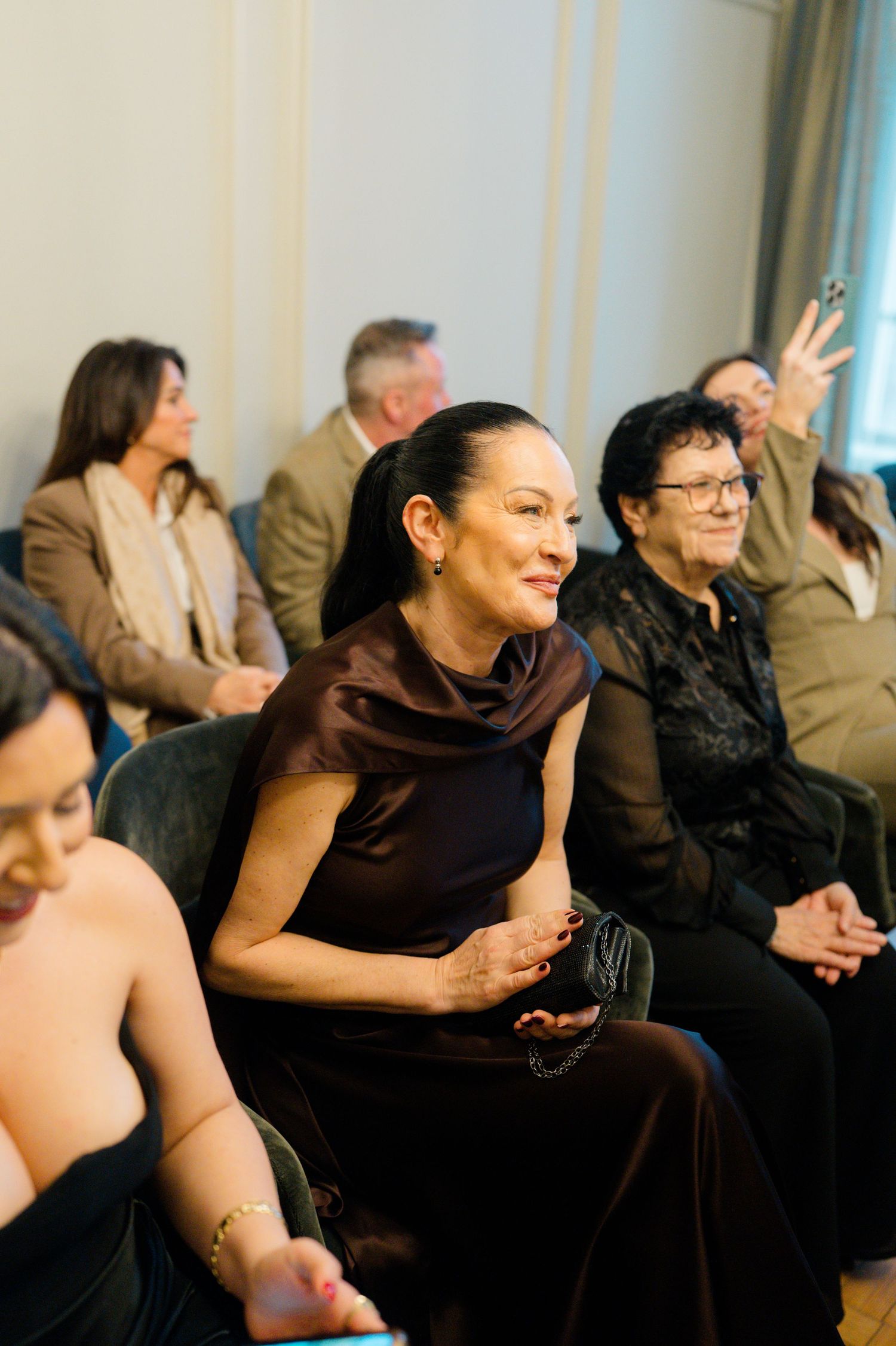 Elegant wedding guests seated at a ceremony, woman in brown satin dress smiling.