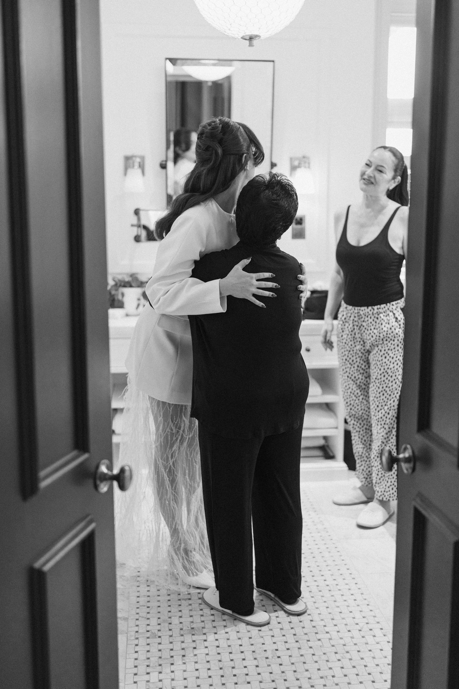 Bride in white feathered wedding outfit sharing an emotional hug with a guest in a hotel bathroom mirror.